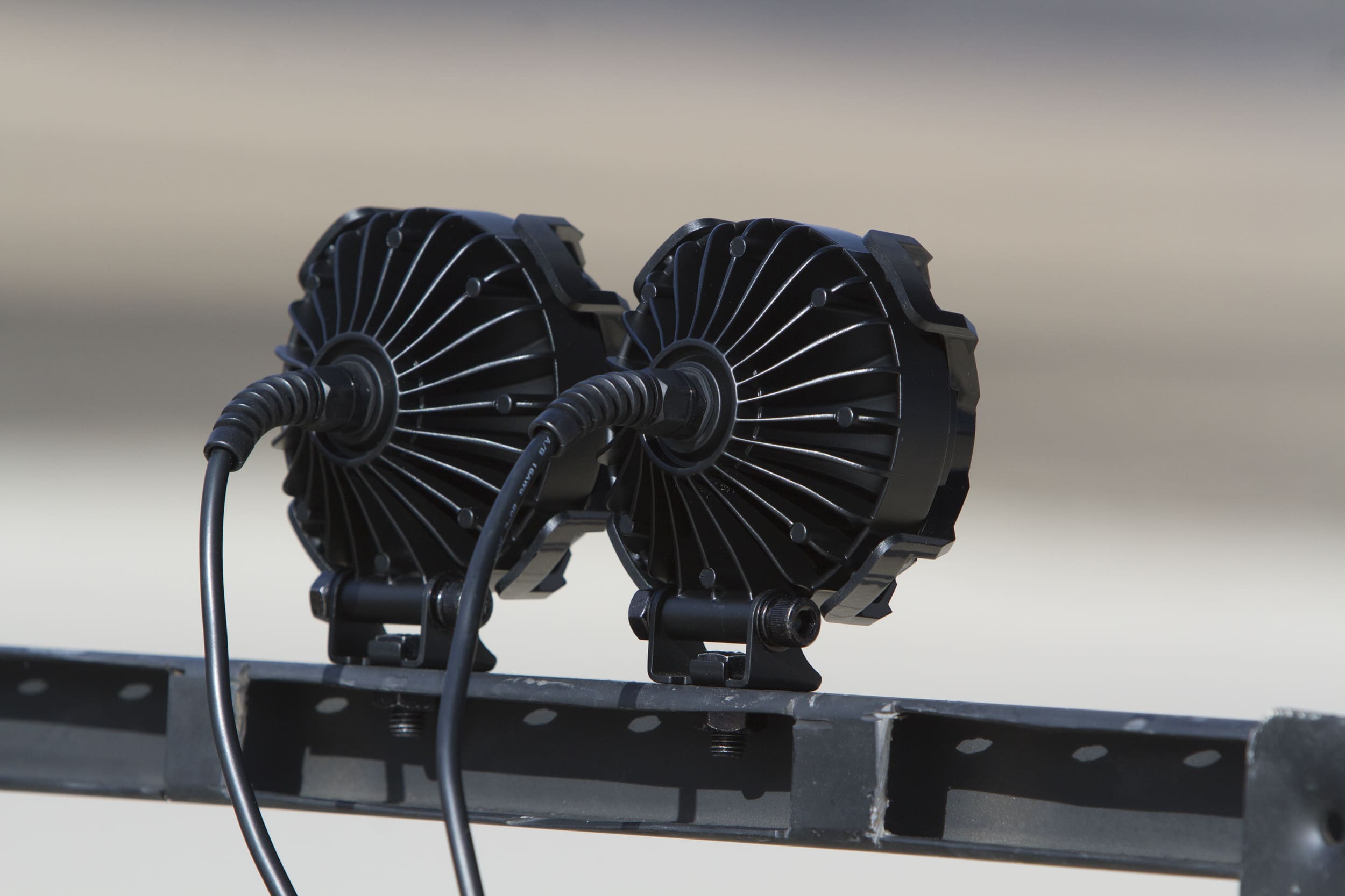 Two black, round electronic devices resembling driving lights, with radiating fins and connected cables, are mounted on a metal bar. The background is blurred and out of focus.