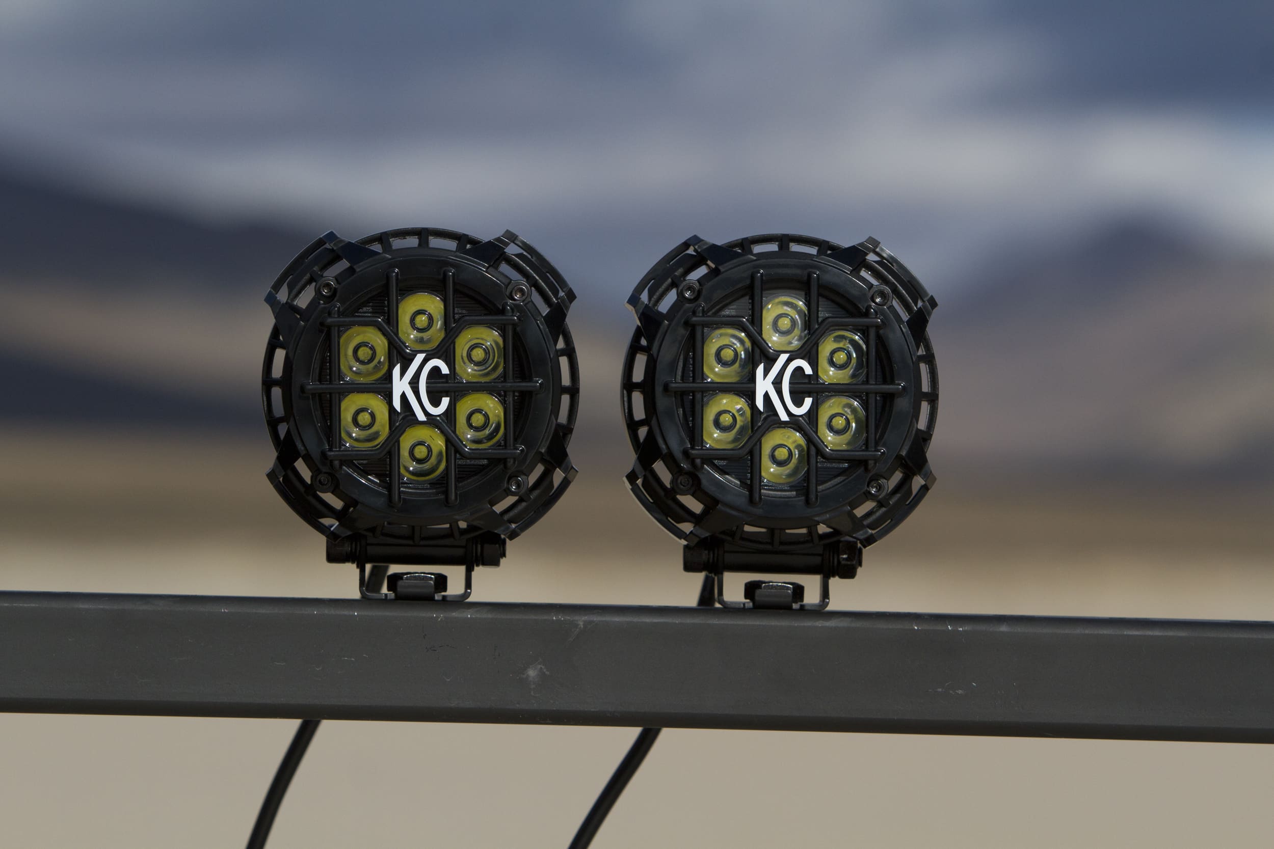 Two round black off-road LED driving lights with KC logos in the center are mounted on a metal bar, with a blurred desert landscape and mountains in the background.