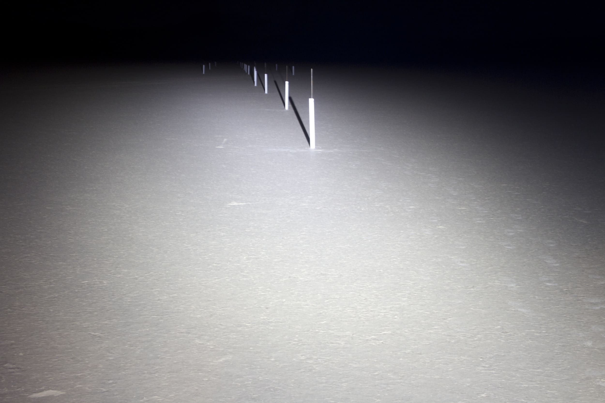A row of white poles extends into the distance across a flat, light-colored surface, illuminated faintly as if by driving lights, before fading into darkness in the background.