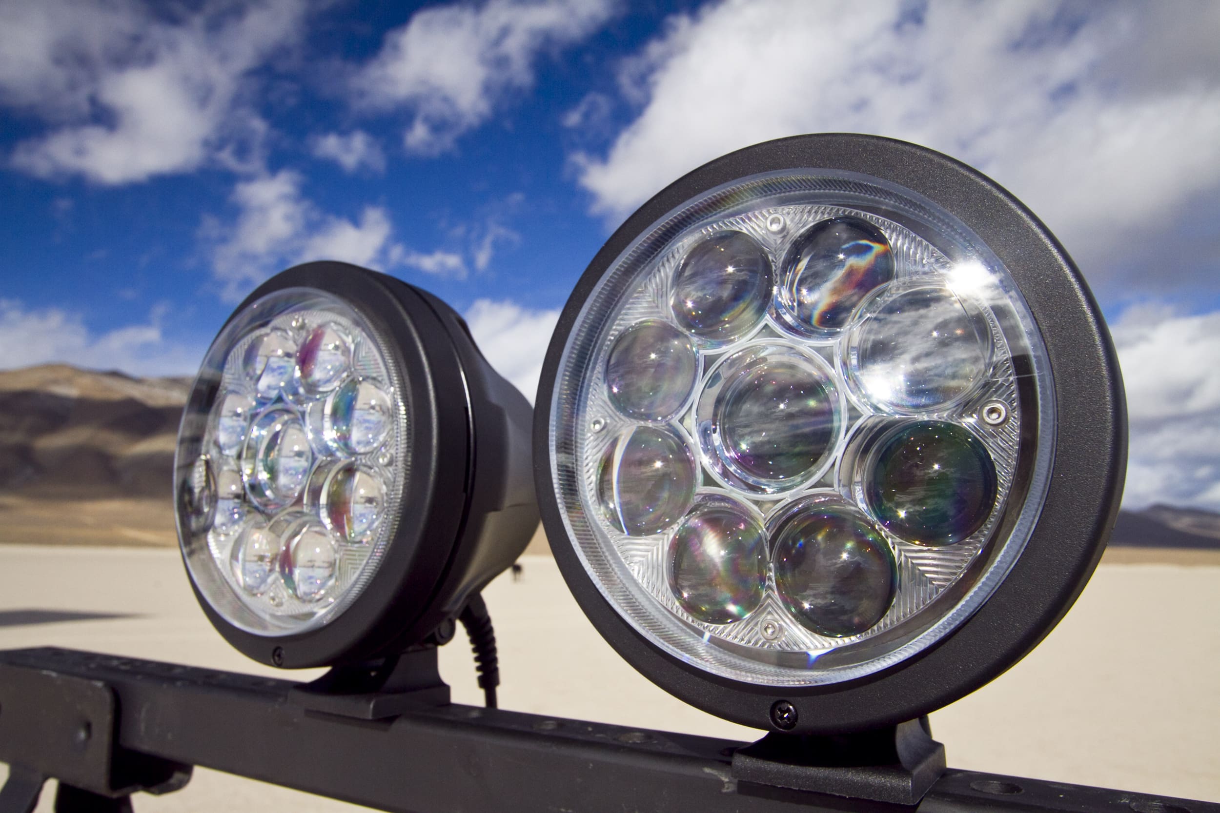 Two round off-road driving lights with multiple LED bulbs are mounted on a metal bar, set against a desert landscape with mountains and a bright blue sky with clouds.