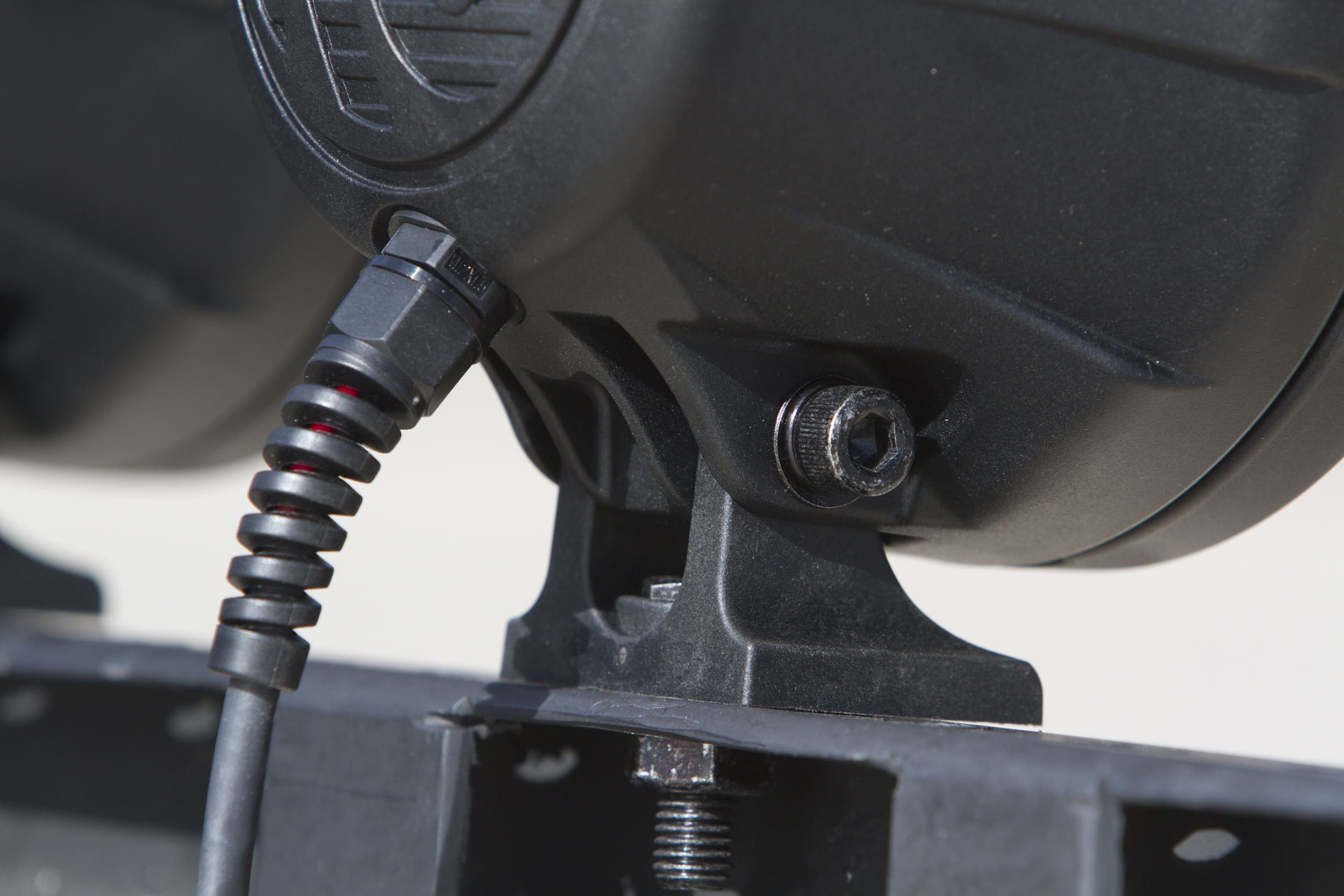 Close-up of a black electronic device with a coiled cable connected and a hex screw securing it to a metal bracket, commonly seen in driving lights setups. The focus is on the connection and mounting details.