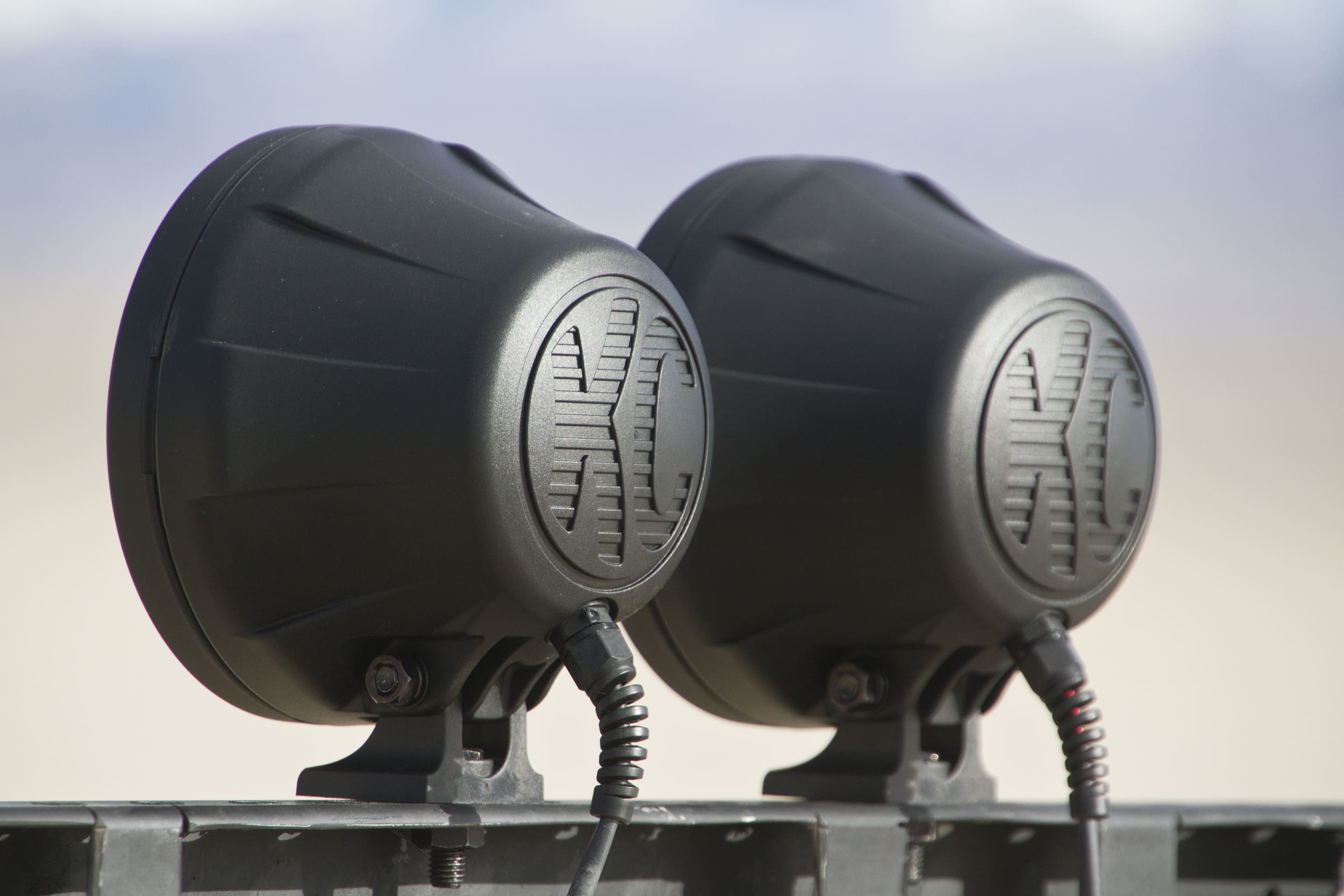 Two black off-road driving lights with KC logos are mounted side by side on a metal bar, with mountains and sky blurred in the background.