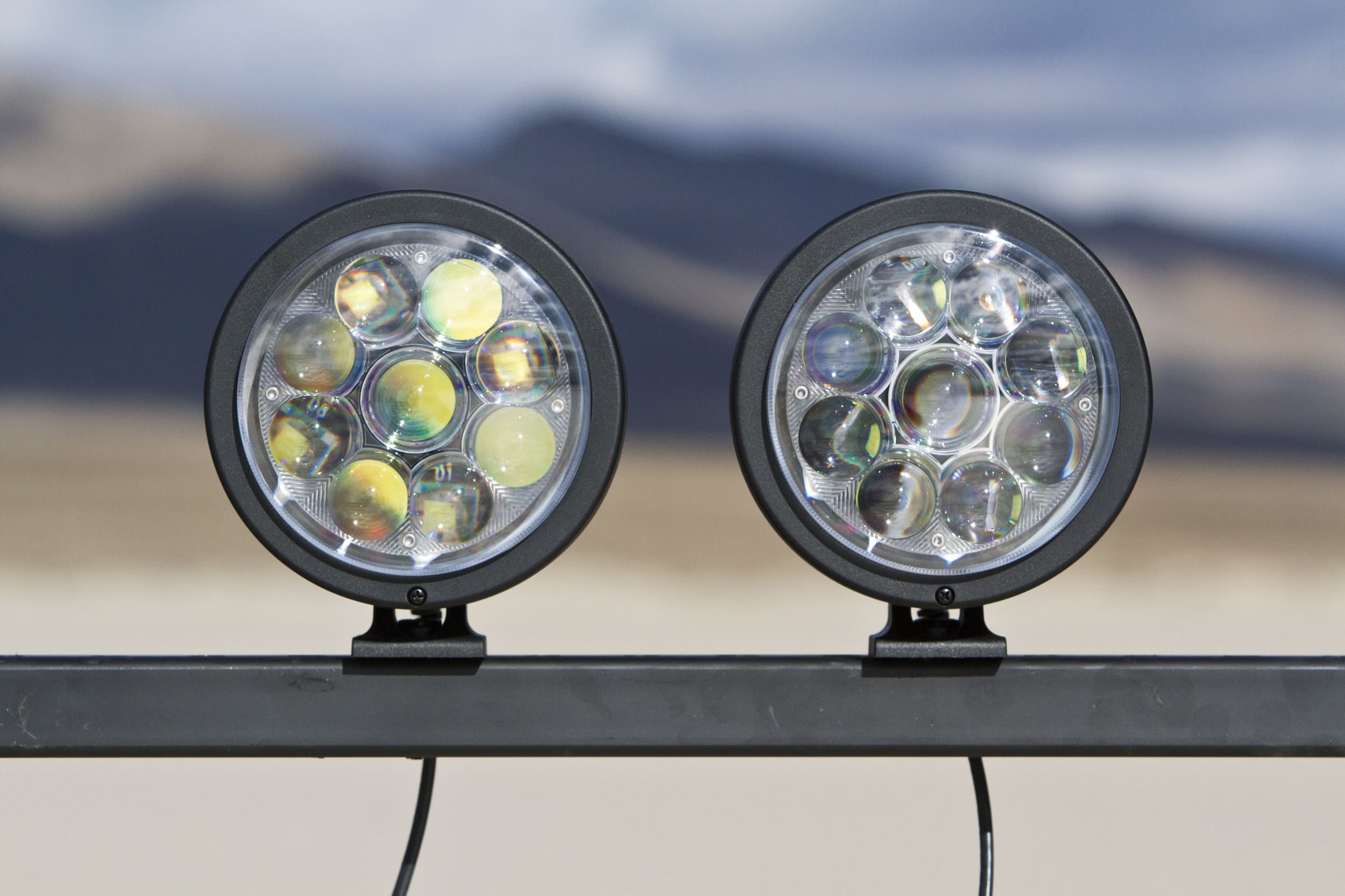 Two round LED driving lights mounted on a horizontal metal bar outdoors, with a blurred mountainous landscape in the background.
