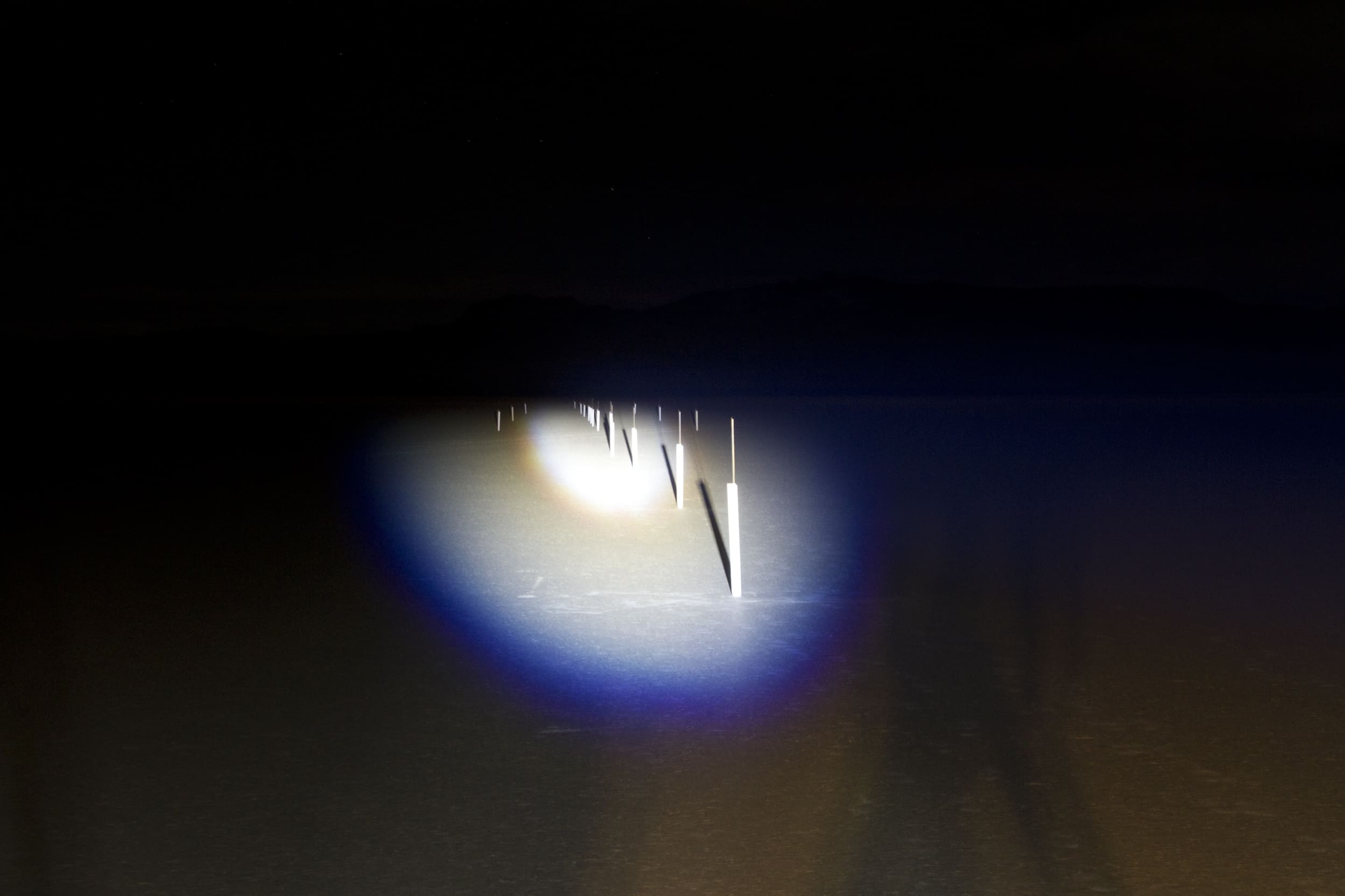 A dark road at night illuminated by driving lights, revealing reflective markers or poles along the center, with a rainbow-like halo effect in the beam.