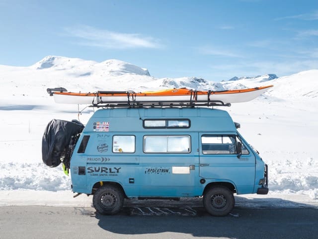 A blue van belonging to Jack Mac, adorned with travel stickers and a kayak on the roof, is parked on a snowy road, with snow-covered mountains in the background under a clear sky.