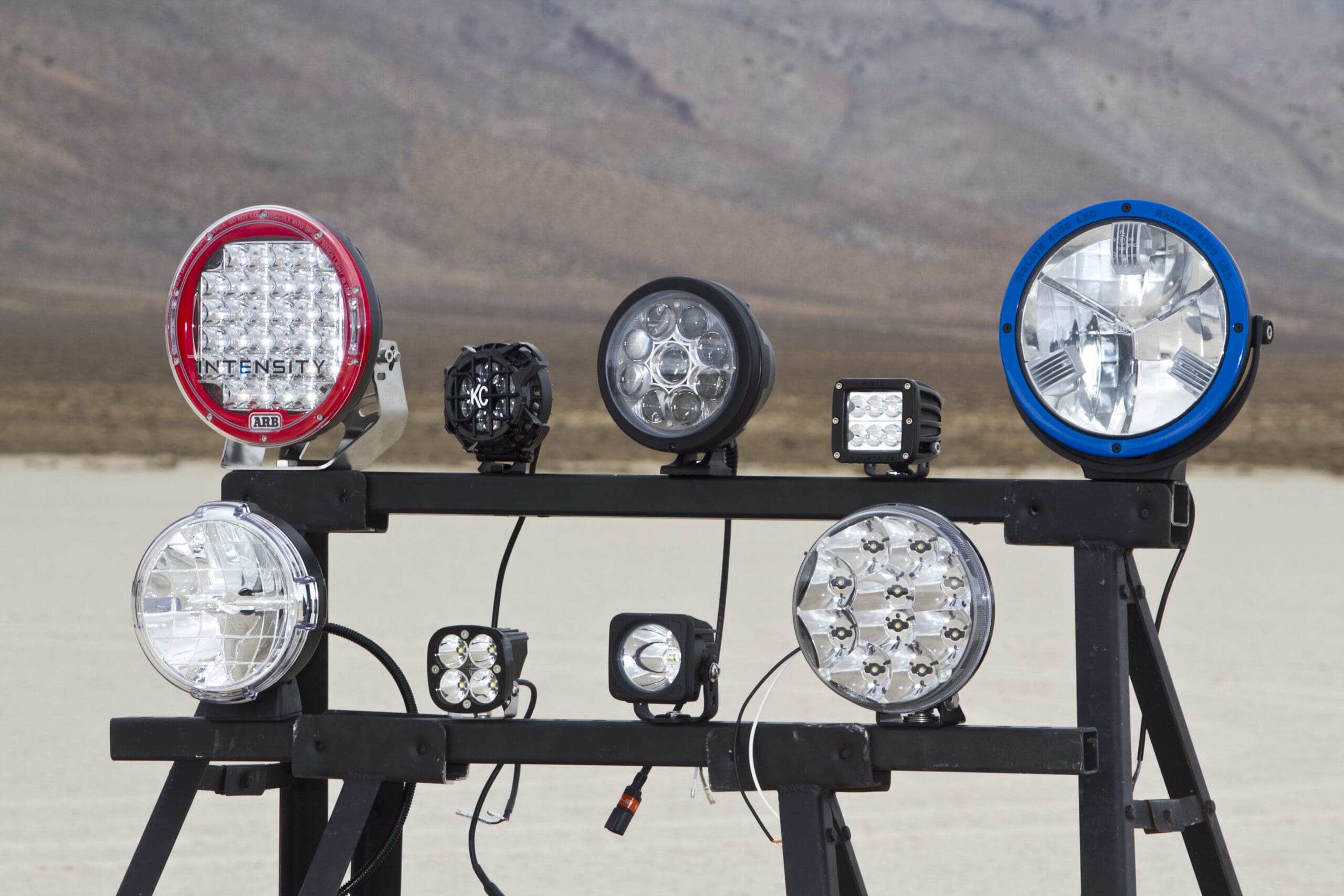 Eight different off-road driving lights are mounted on a black metal frame, displayed outdoors on a sandy, desert-like plain with rocky hills in the background.