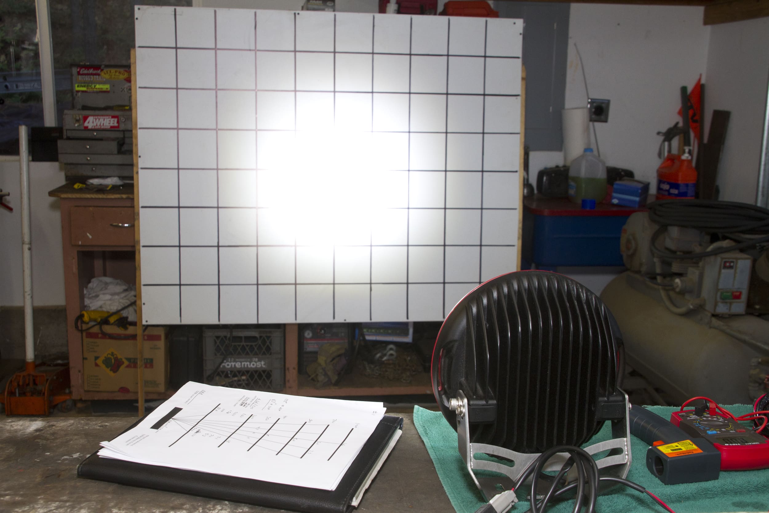 A bright light, reminiscent of powerful driving lights, shines on a grid-covered board in a workshop. A light meter, clipboard, and other tools are on a workbench in the foreground.