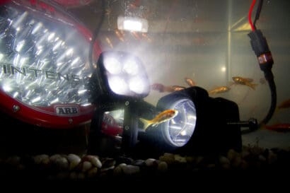 Goldfish swim in a tank illuminated by large underwater spotlights, a red ARB light, and even submerged driving lights that cast sharp beams through the water. Pebbles cover the tank floor, and wires are visible winding between them.