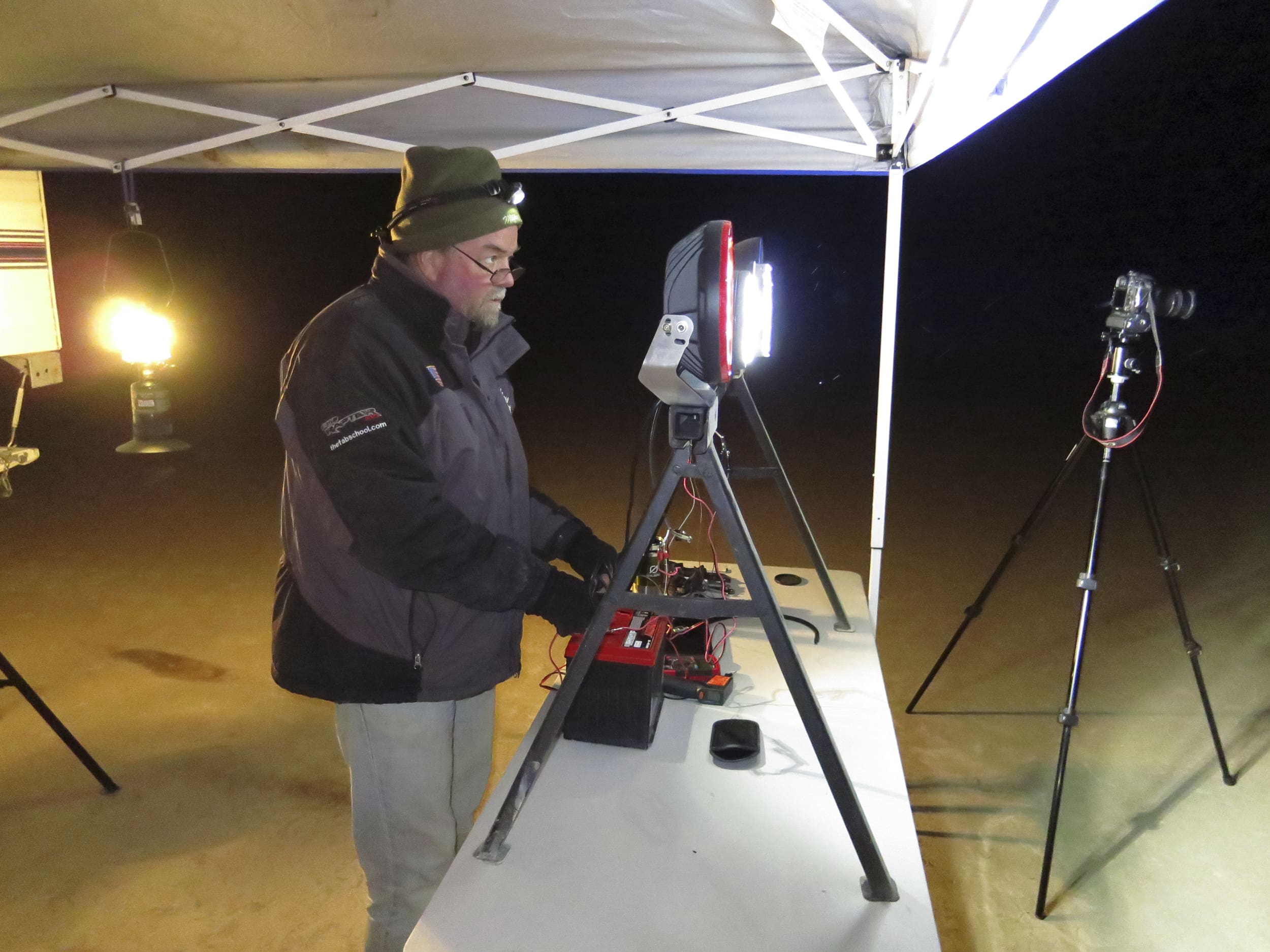 A person in a coat and beanie works at a lit table with electronic equipment under a canopy at night. A lantern, tripod, and camera are also visible on the scene, with the soft glow of driving lights illuminating the area.