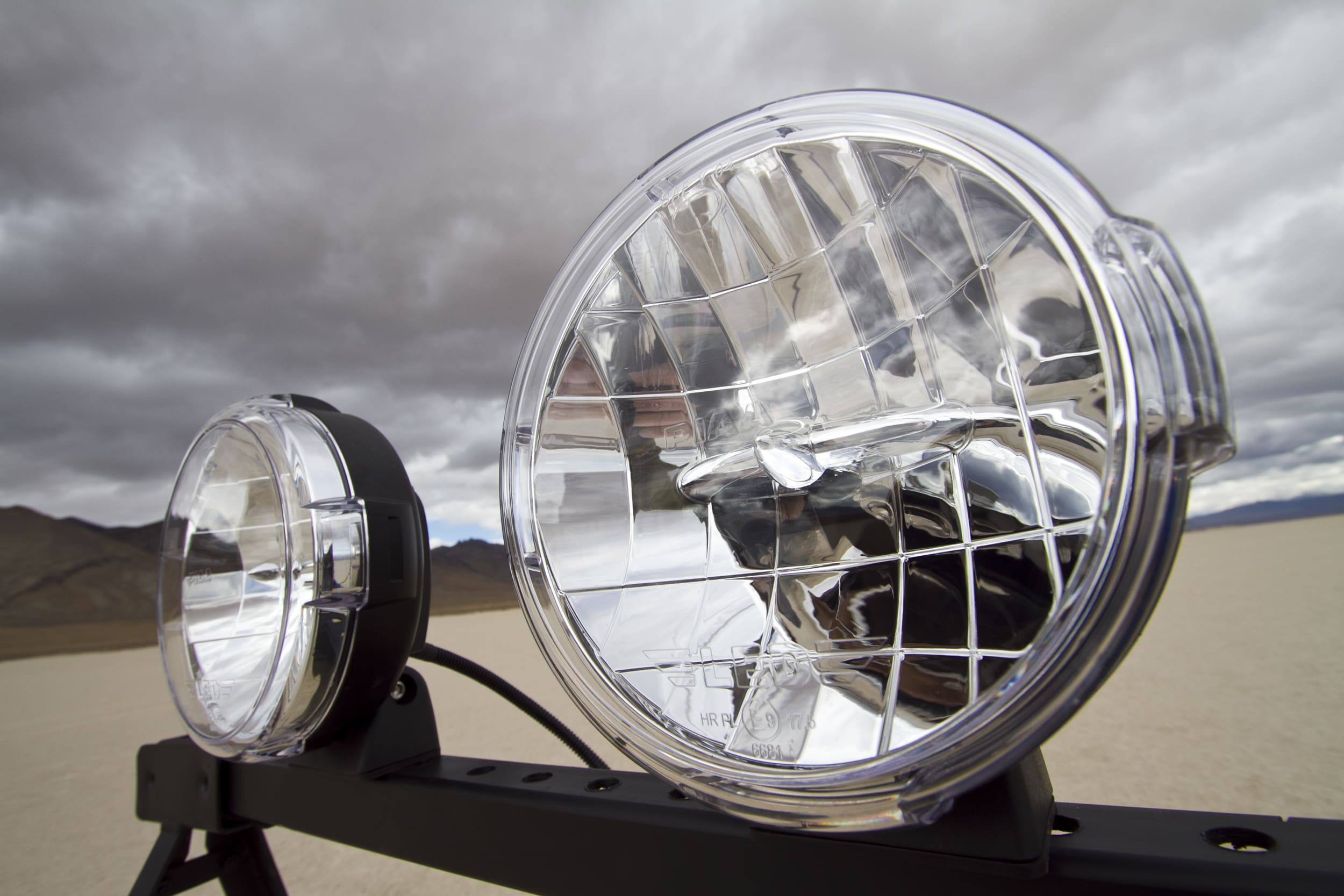 Close-up of two round driving lights mounted on a bar, with a cloudy sky and barren landscape in the background.