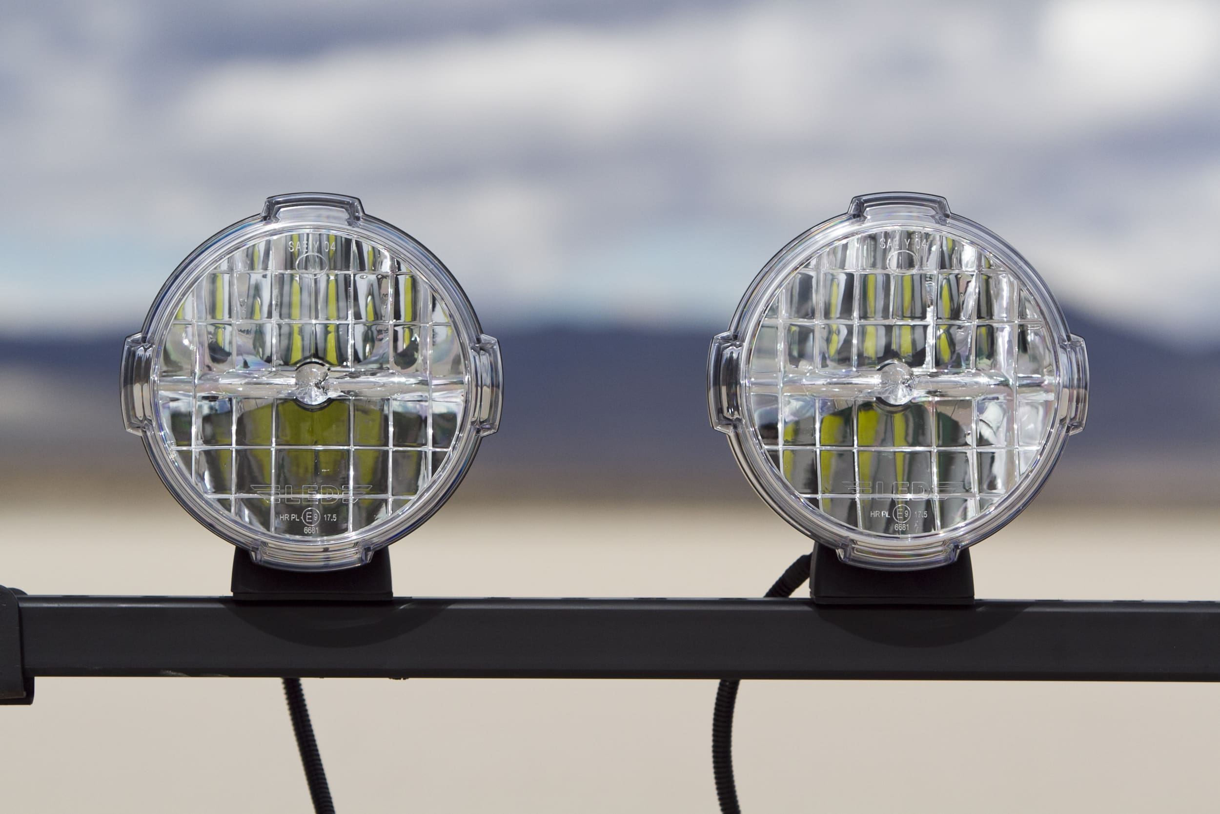 Two round, clear glass driving lights with grid covers are mounted side by side on a black metal bar. The background is blurred, showing an open landscape with mountains and a cloudy sky.