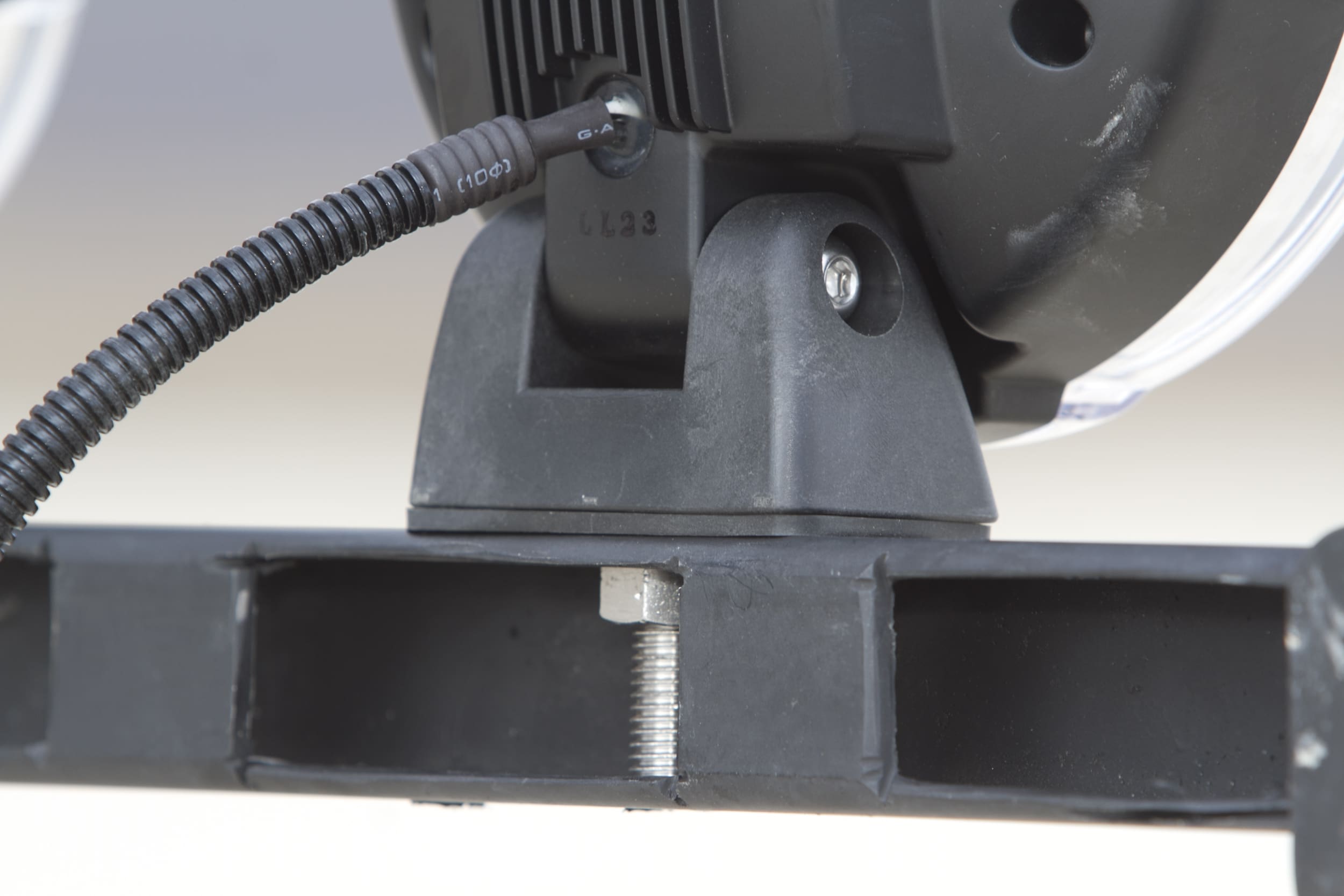 Close-up of a black mounting bracket secured with a bolt and nut, attached to a device—such as driving lights—with a visible wire and screw. The background is blurred.