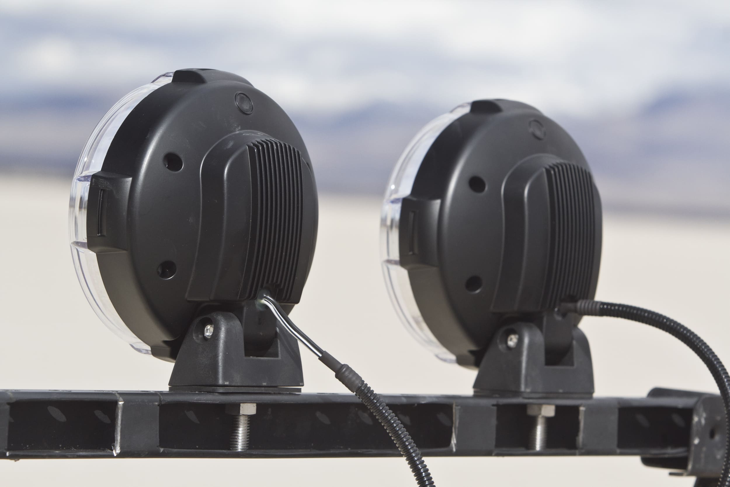 Two round black driving lights are mounted on a metal bar, facing away from the camera, with a blurred outdoor landscape in the background.