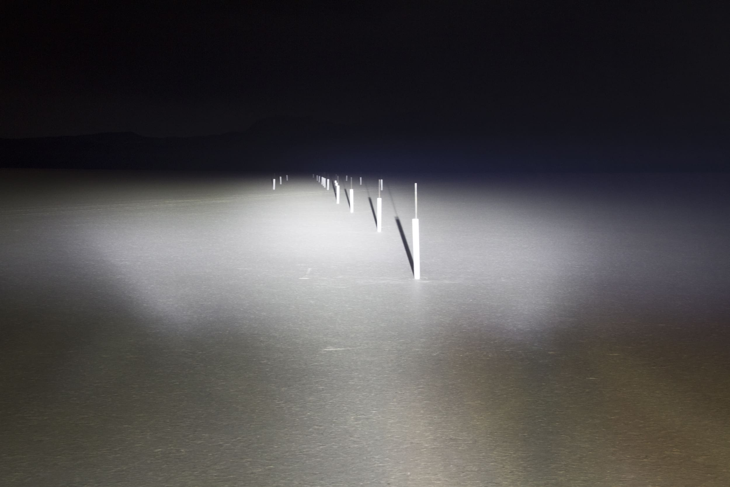 A row of white poles stands in the middle of a dark, flat landscape illuminated by driving lights, fading into darkness in the distance.