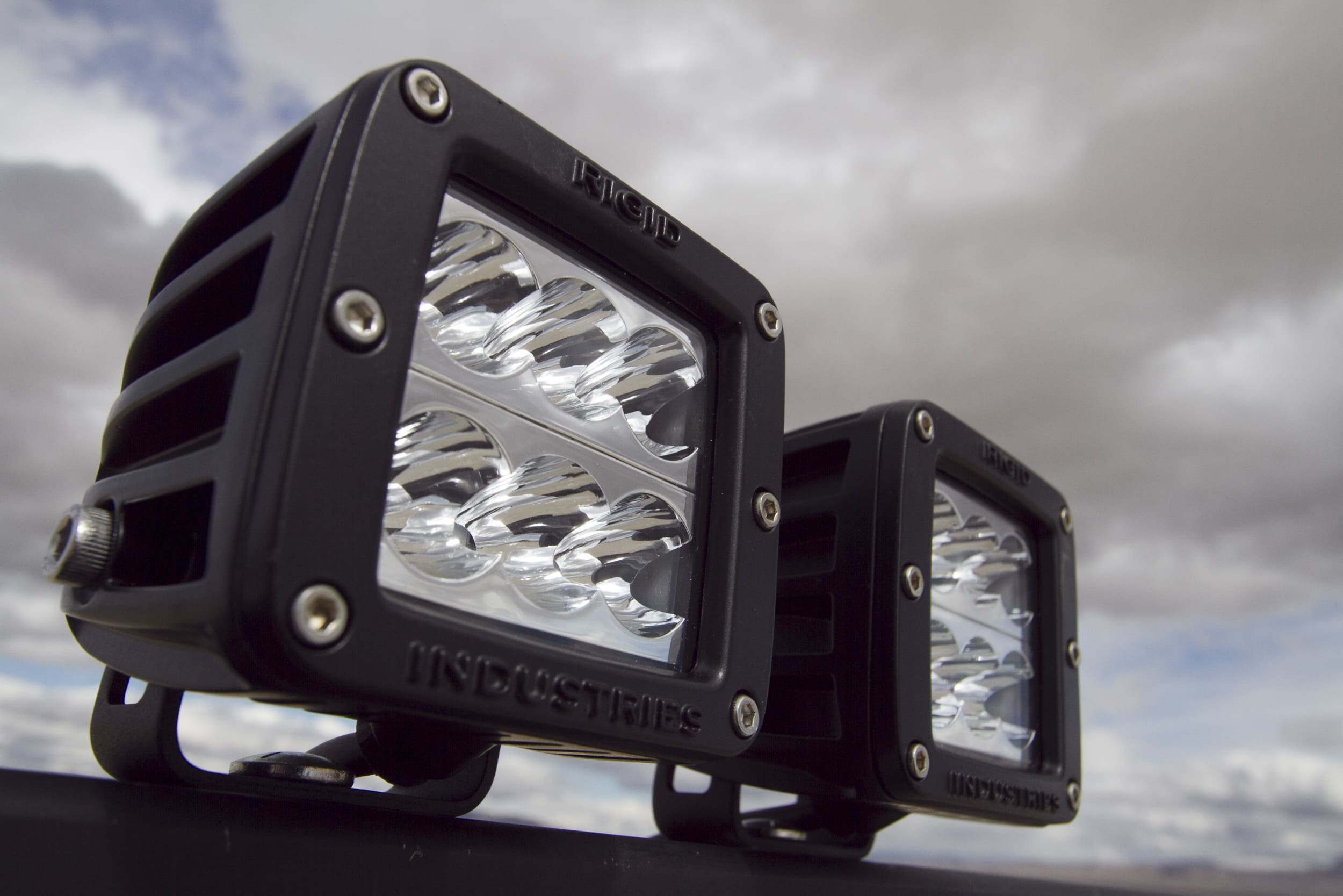 Two black, square-shaped LED driving lights are mounted outdoors against a cloudy sky, with the focus on their clear lenses and sturdy casing.