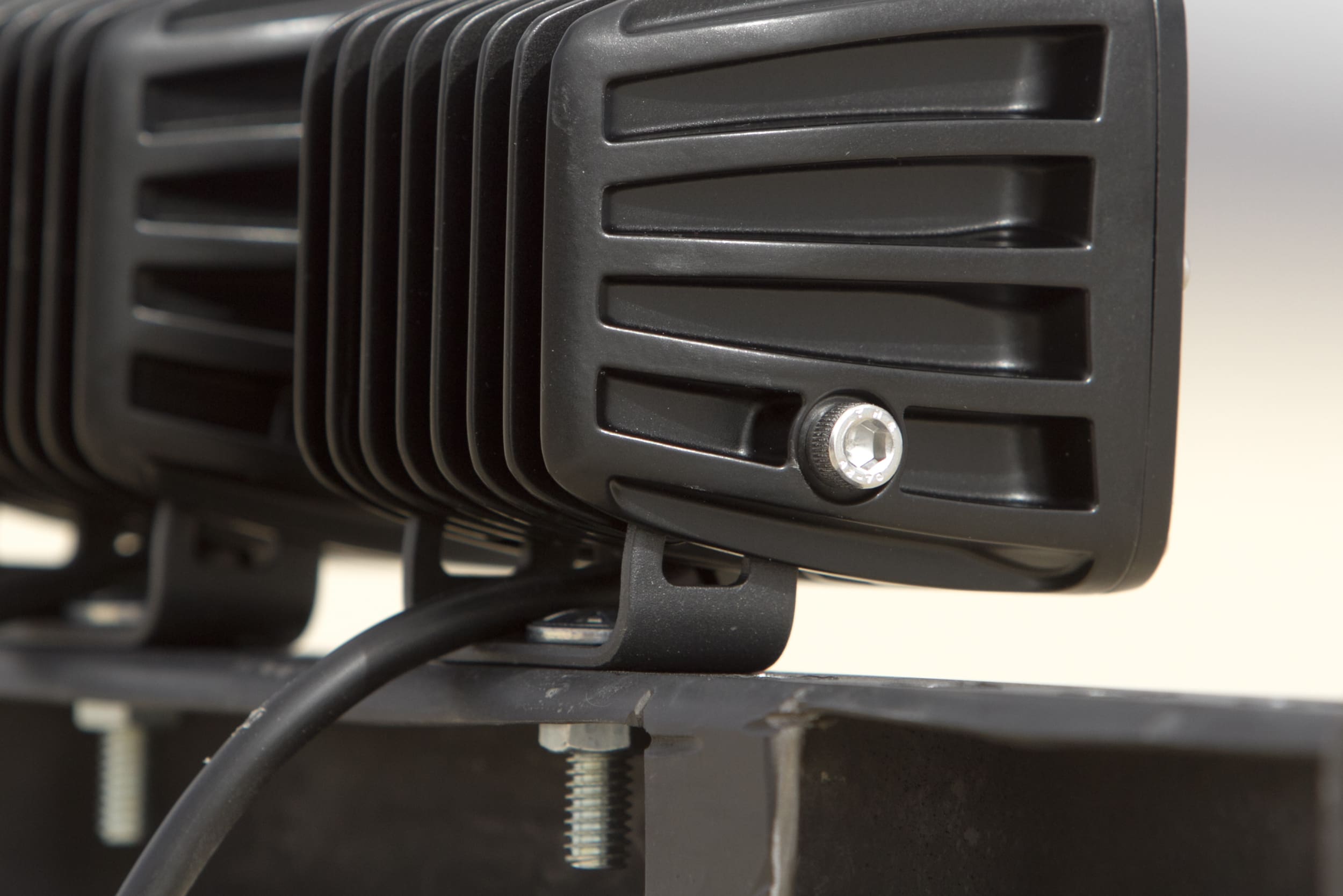 Close-up shot of a black LED light bar mounted on a metal bracket, commonly used as driving lights. The ribbed heat sink design and a single hexagonal screw are visible on the side of the light housing. A power cable runs from the light to the mounting base below.