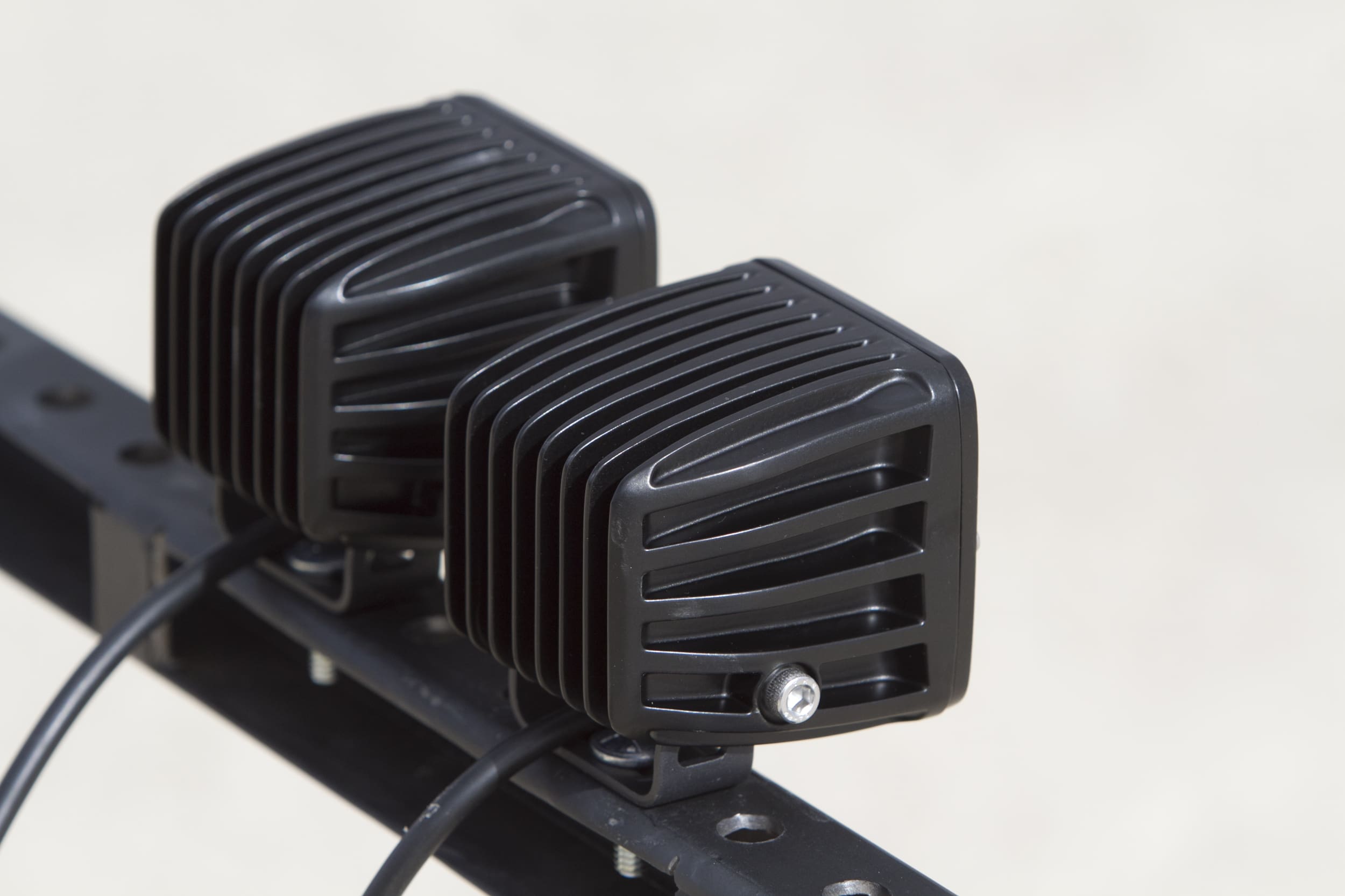 Two black, rectangular LED driving lights with finned heatsinks are mounted side by side on a metal bar, with a visible screw and power cables attached to each light. The background is blurred and neutral.