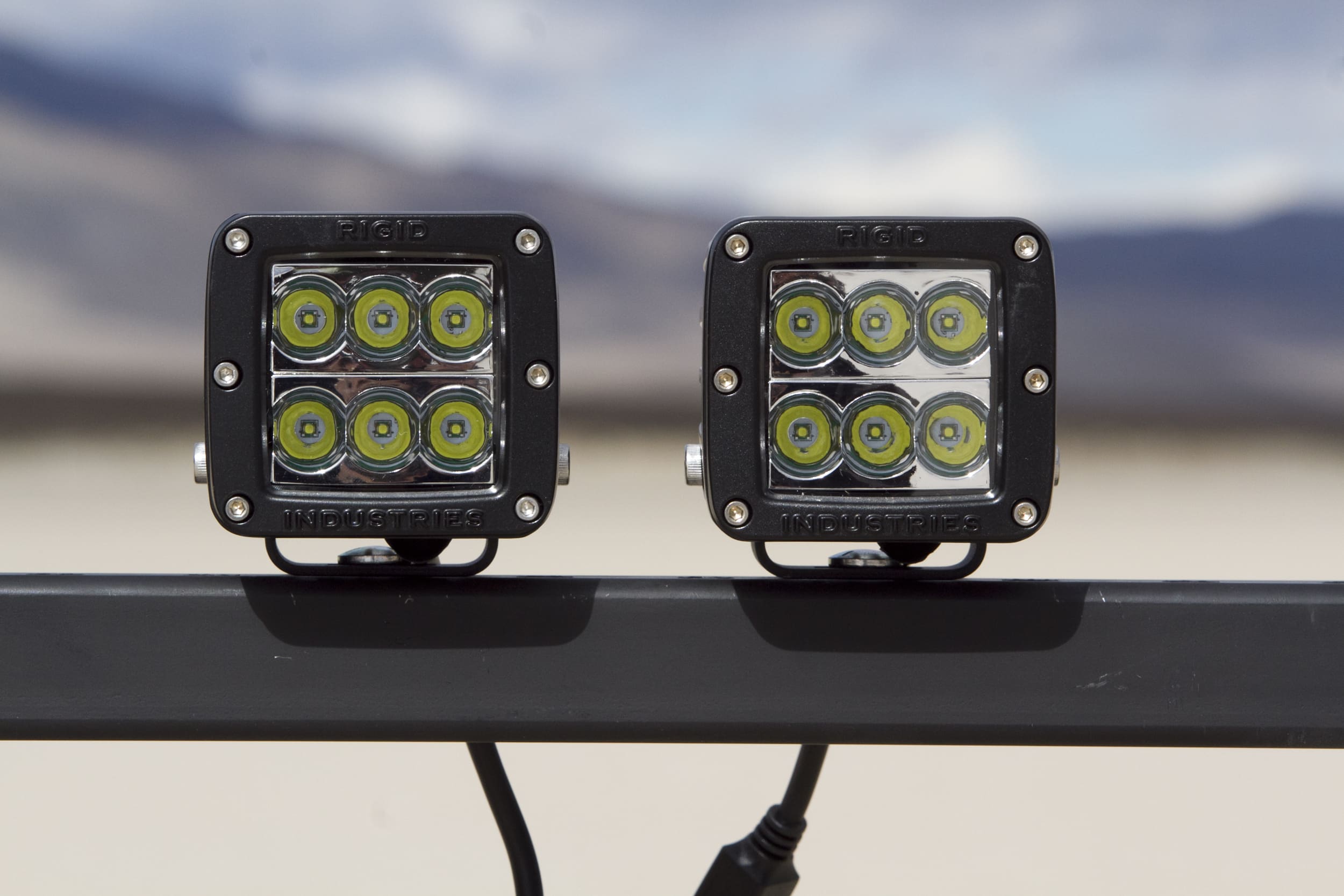 Two square LED driving lights pods with six bulbs each mounted on a metal bar outdoors, with a blurred desert landscape in the background.