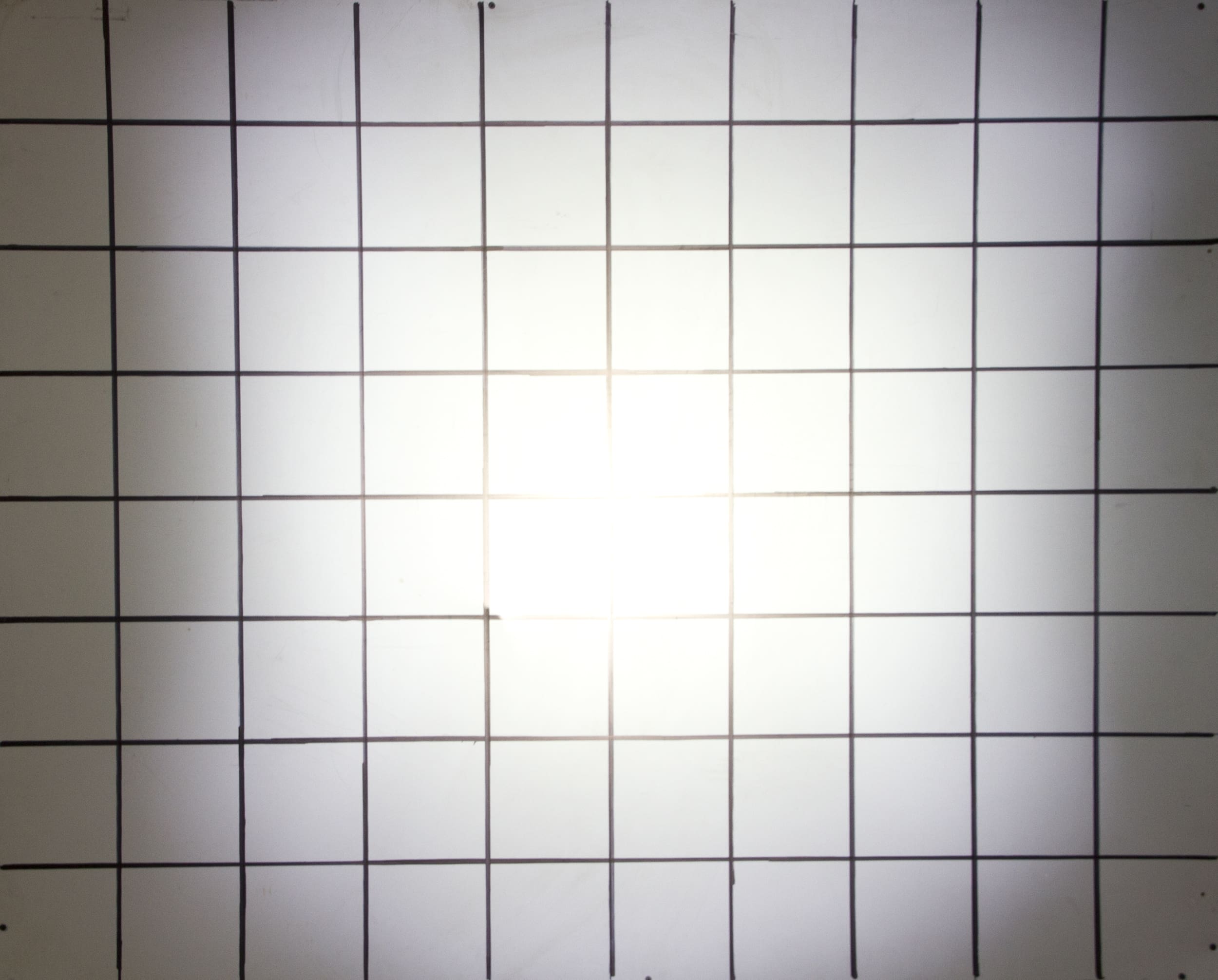 A white surface with evenly spaced black grid lines forming squares. The center appears brighter, creating a light gradient effect toward the edges, reminiscent of the focused illumination produced by driving lights.