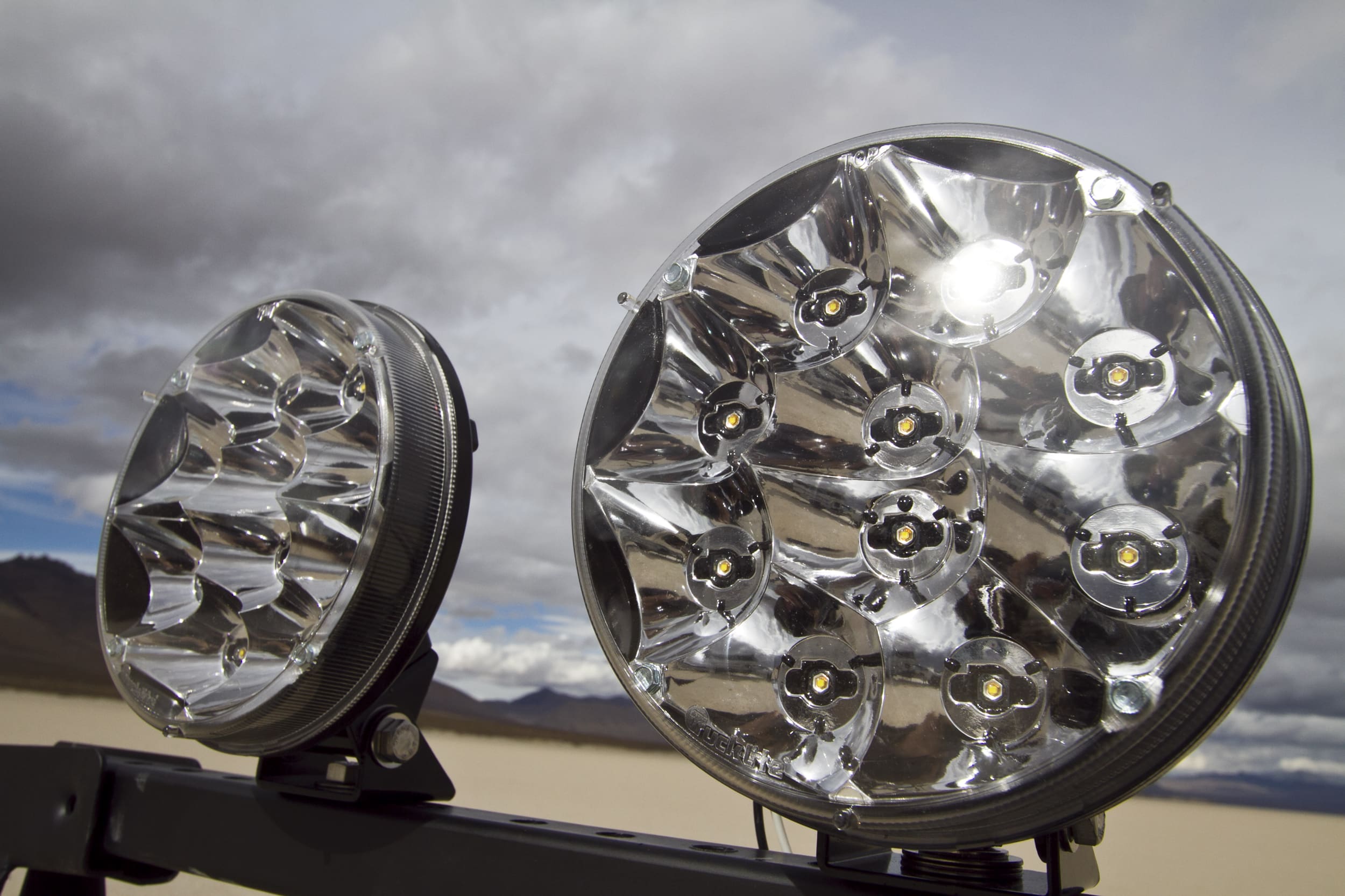 Close-up of two round, multi-LED driving lights mounted on a vehicle with a cloudy sky and distant mountains in the background.