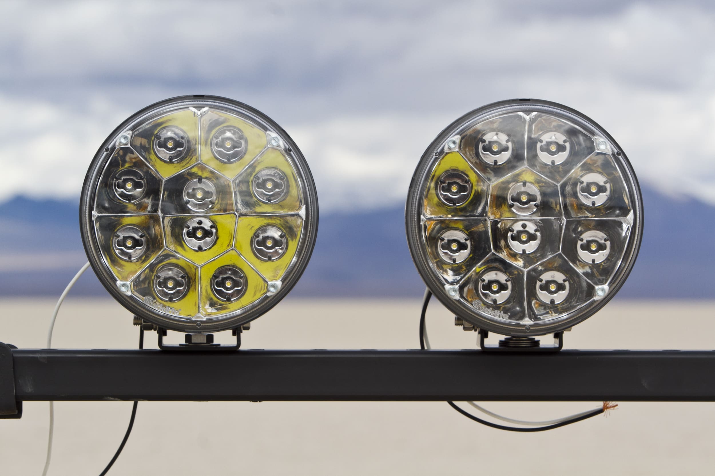 Two round, multi-LED driving lights are mounted side by side on a metal bar, with a desert landscape and mountains blurred in the background.