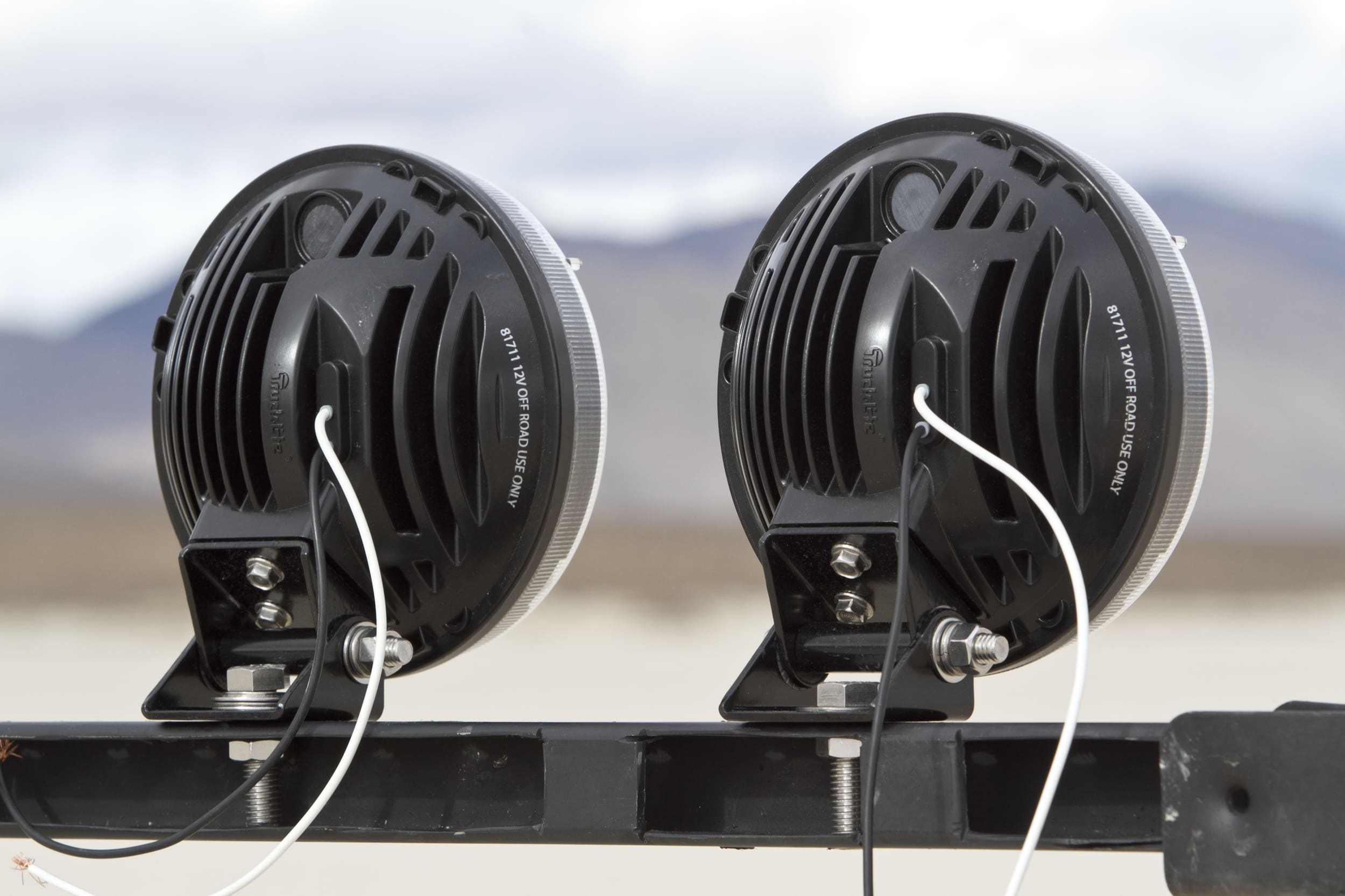 Two round black driving lights with wiring are mounted on a metal bar, facing away from the camera. A blurred landscape and mountains are visible in the background.