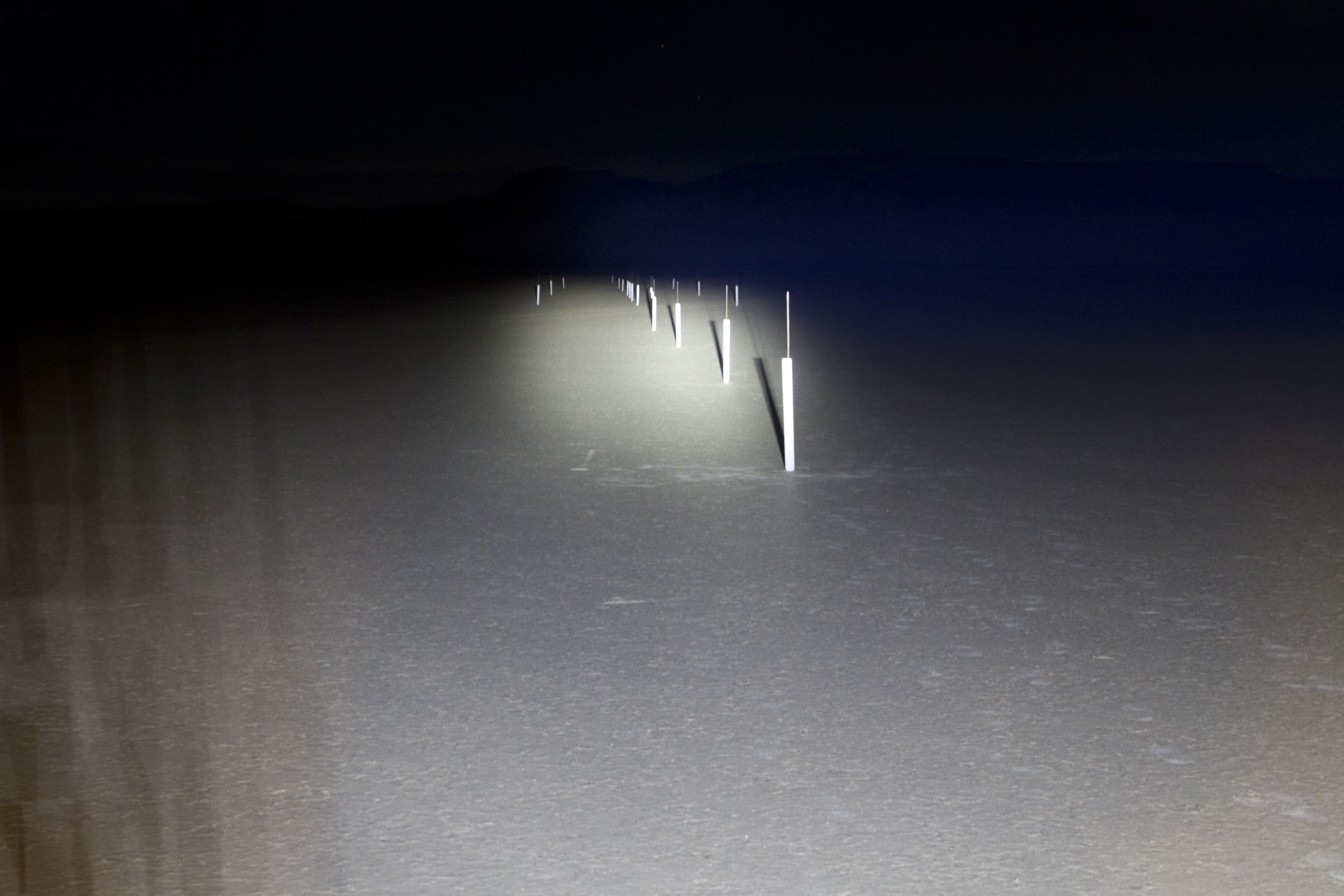 Headlights and driving lights illuminate a row of white posts stretching into the darkness across a flat, barren landscape at night.
