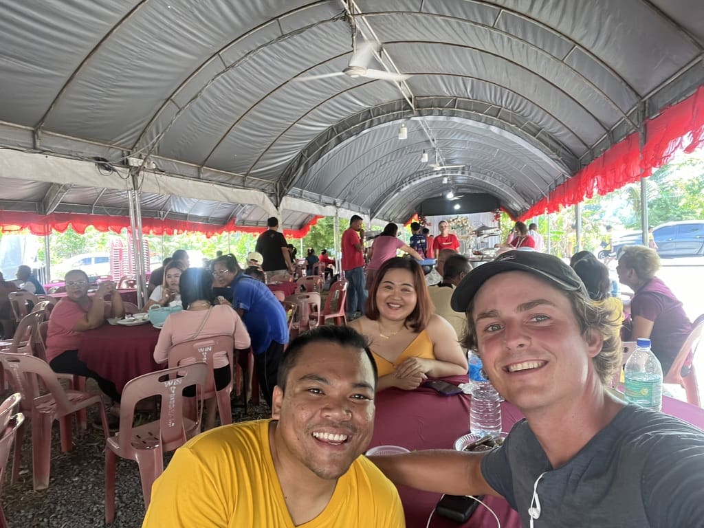 Two smiling men take a selfie at an outdoor event under a large tent. People sit at round tables covered with pink tablecloths, eating and socializing. Bright daylight and festive atmosphere.