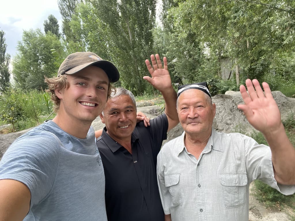 Three men stand outdoors smiling and waving at the camera, with trees and rocks in the background. The man on the left wears a cap, the others wear collared shirts. It is a bright, green, outdoor setting.