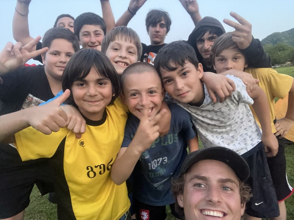 A group of smiling kids and a young adult take a close-up selfie outdoors on a field, some giving thumbs up and peace signs.