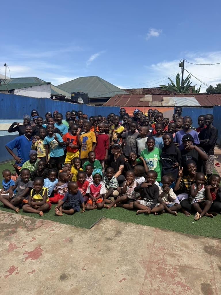 A large group of children and adults pose and smile together outside on a sunny day, with a blue wall and rooftops in the background.