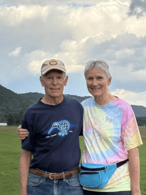 An older man and woman stand together outdoors on grass, smiling at the camera during a Celebration of Life. He wears a navy T-shirt and beige hat; she wears a tie-dye shirt and blue fanny pack. Cloudy sky and hills are in the background, capturing a warm moment shared by Arden Kysely and his companion.