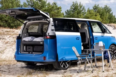 A blue and white VW electric campervan is parked on sand with its rear hatch and side door open, revealing cozy bedding inside. Nearby are two chairs and a small table, set up for outdoor leisure. Pine trees are in the background.