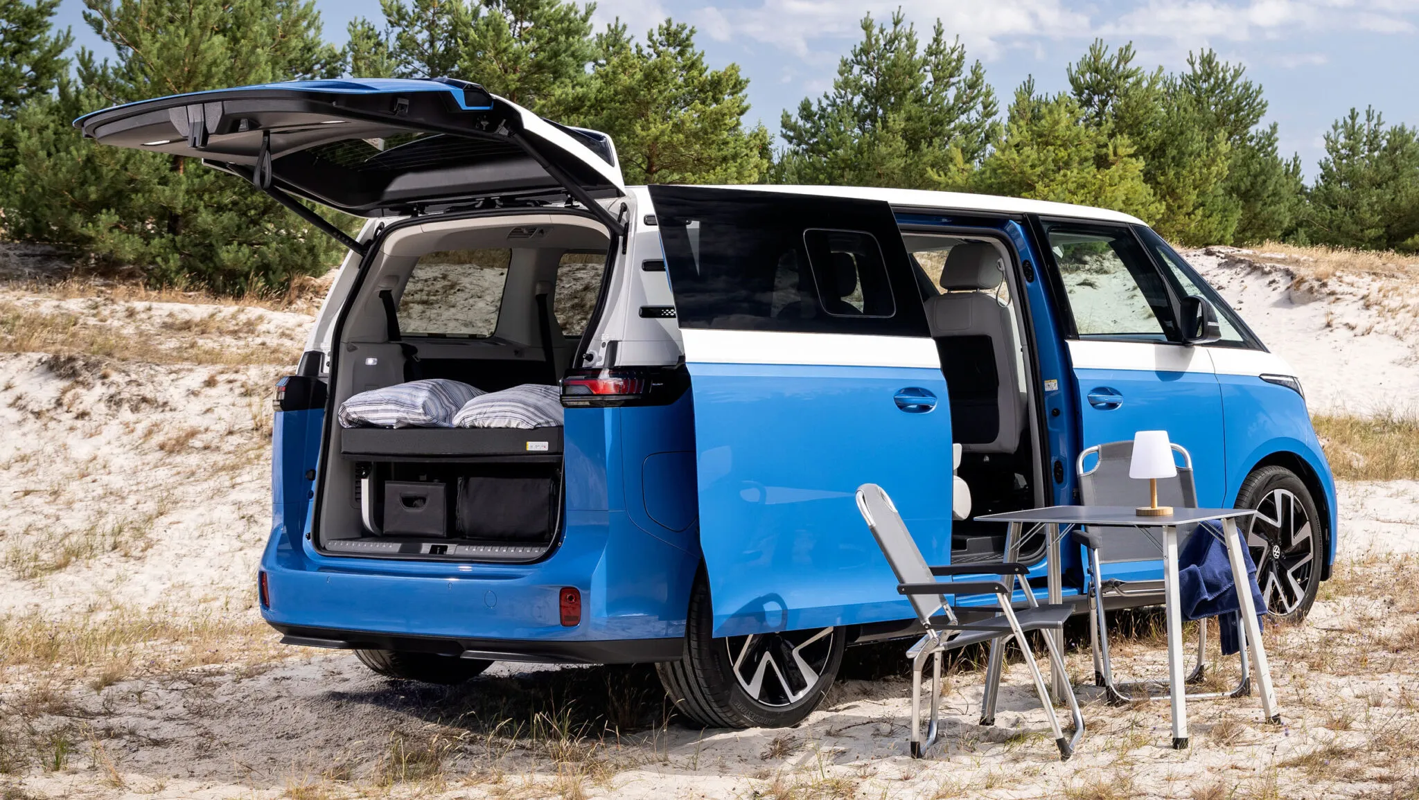 A blue and white VW electric campervan is parked on sand with its rear hatch and side door open, revealing cozy bedding inside. Nearby are two chairs and a small table, set up for outdoor leisure. Pine trees are in the background.