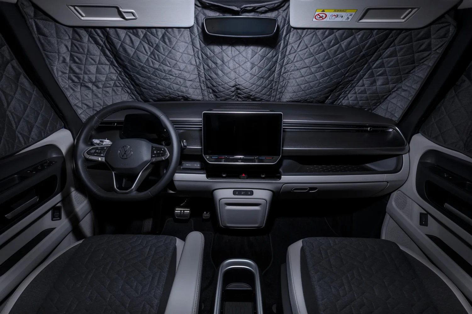 Interior view of a modern VW electric California campervan with quilted privacy shades covering the windshield and windows, a central touchscreen, two seats, and a minimalist dashboard design.