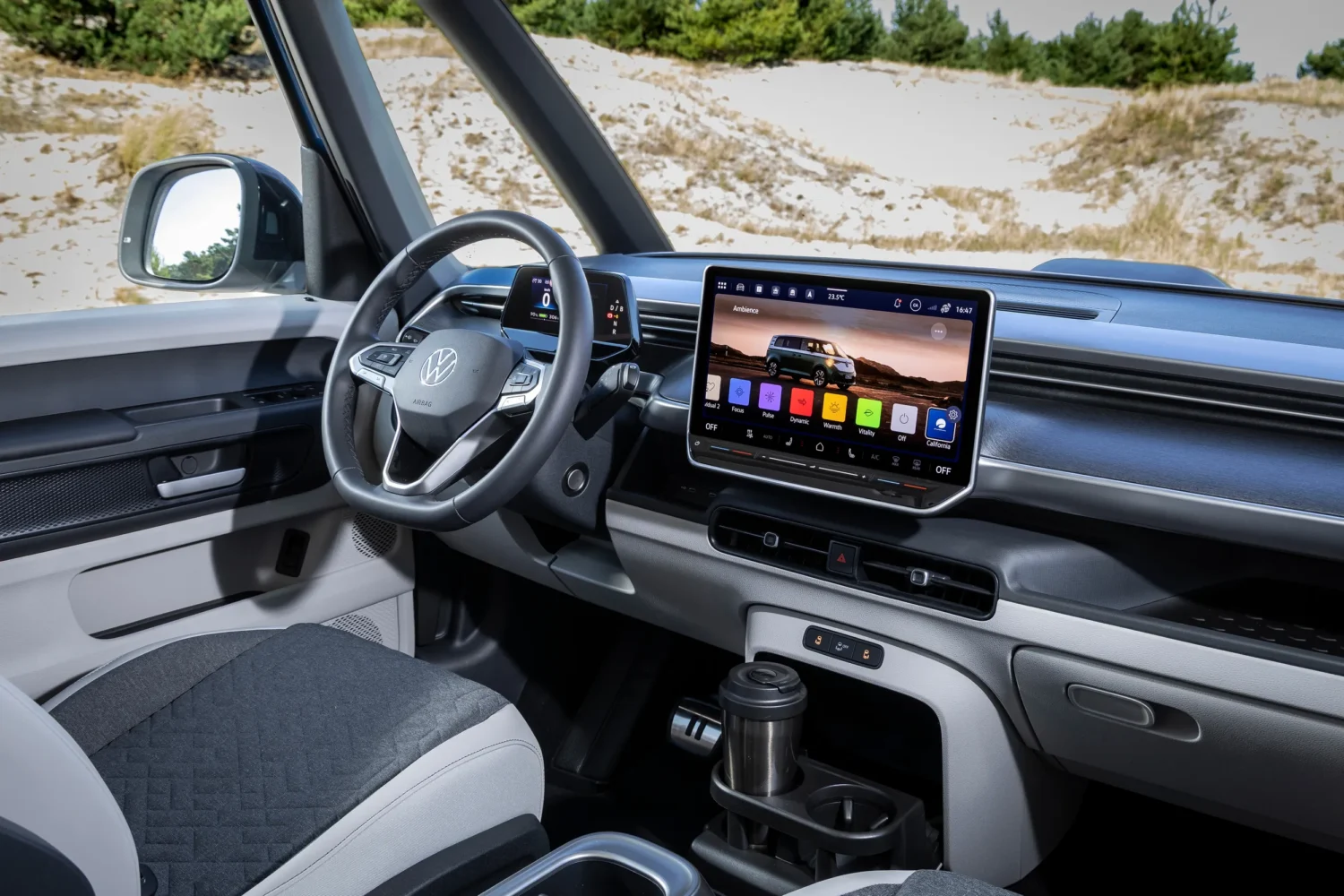 Interior of a Volkswagen vehicle showing the steering wheel, dashboard, large touchscreen display with apps, and gear selector—hinting at the innovative comforts of a fully electric VW California campervan—with a sandy outdoor scene visible through the window.
