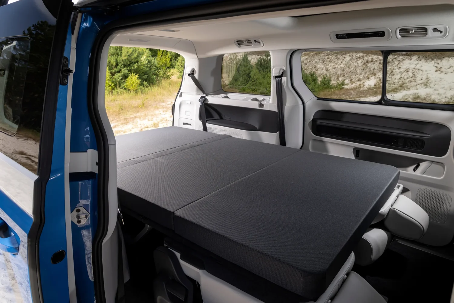 The rear seats of a blue, fully electric VW California are folded flat, forming a spacious bed. Sunlight streams through large windows, and a green, wooded area is visible outside—perfect for relaxing in your electric campervan.