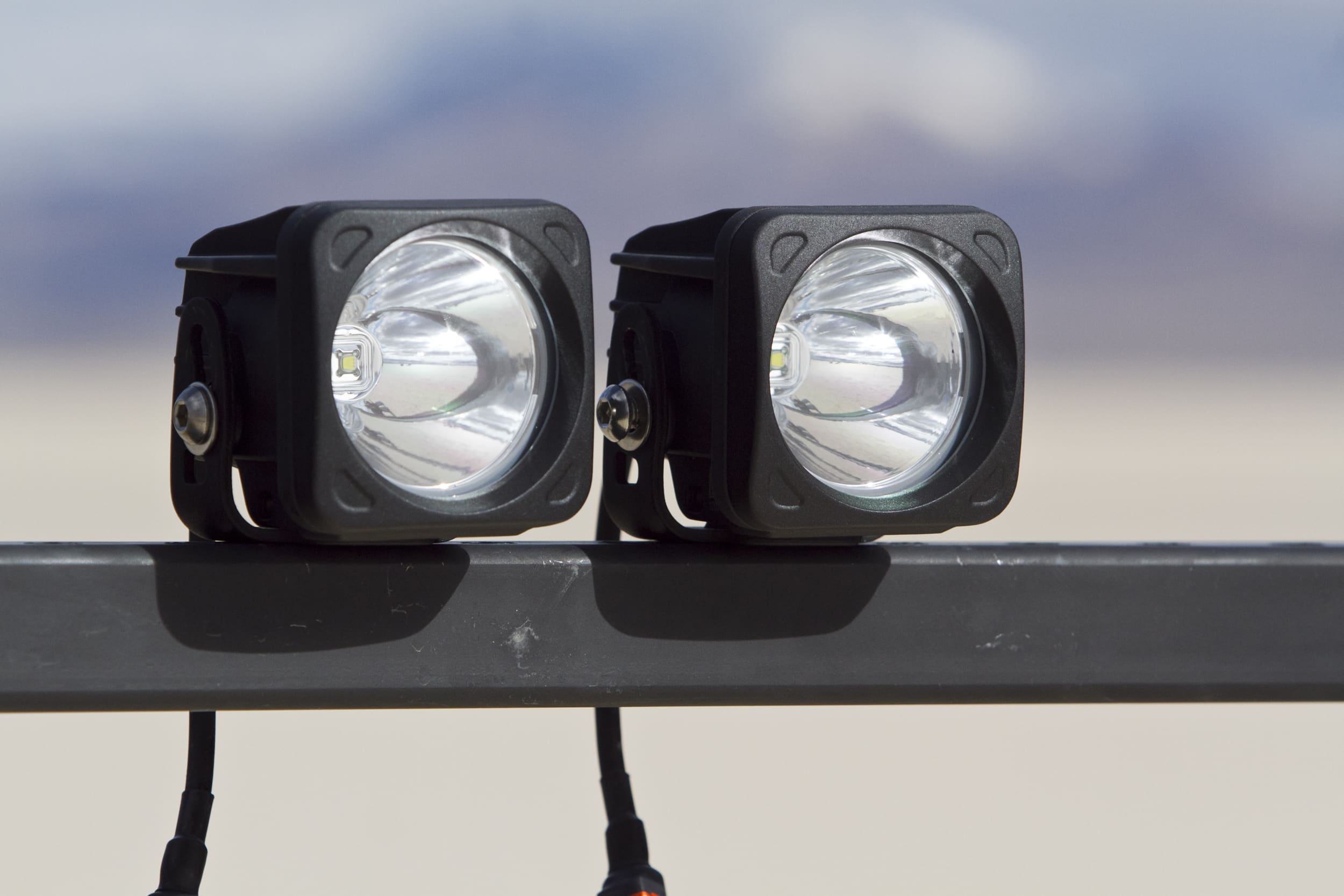 Two square-shaped LED driving lights are mounted side by side on a horizontal metal bar, with blurred mountains and sky in the background.