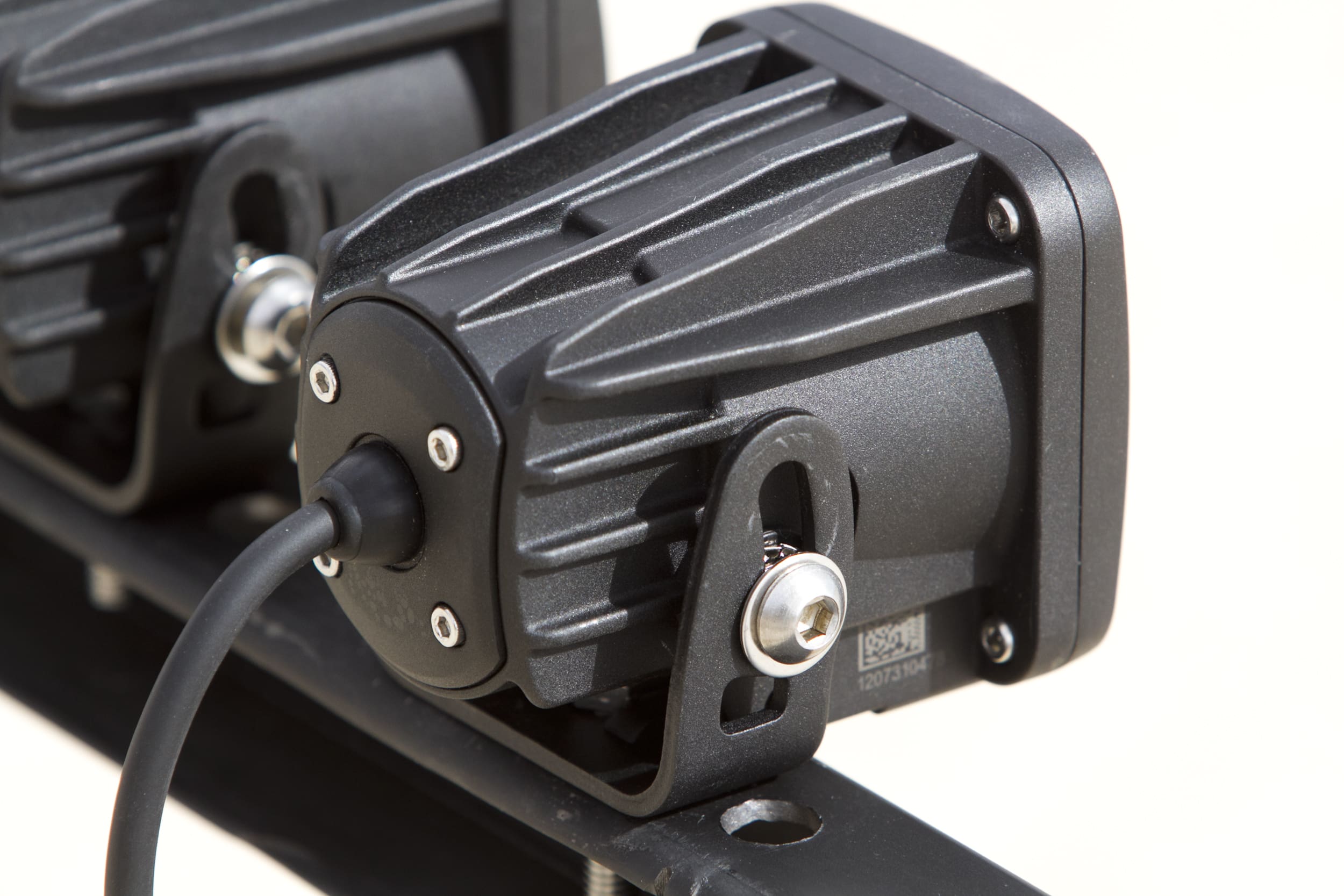 Close-up of a black, mounted LED driving light or spotlight with a cable attached, showing its textured housing and mounting bracket secured with bolts. Another similar unit is partially visible in the background.
