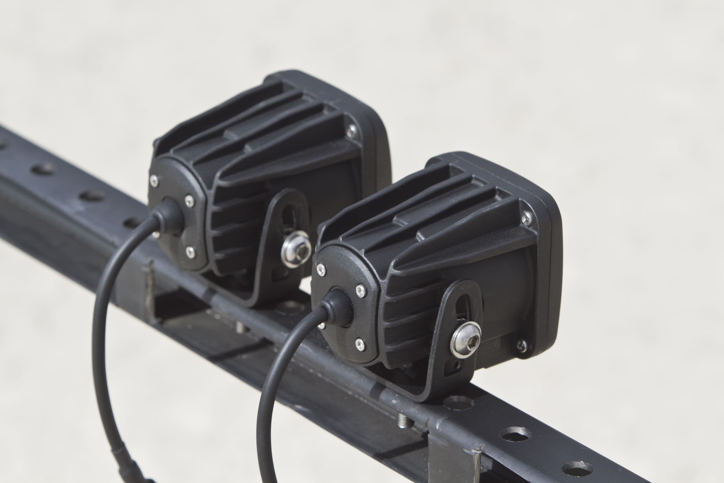 Two black rectangular electronic devices with cables attached are mounted side by side on a metal rail against a plain light background, resembling compact control units often used for driving lights.