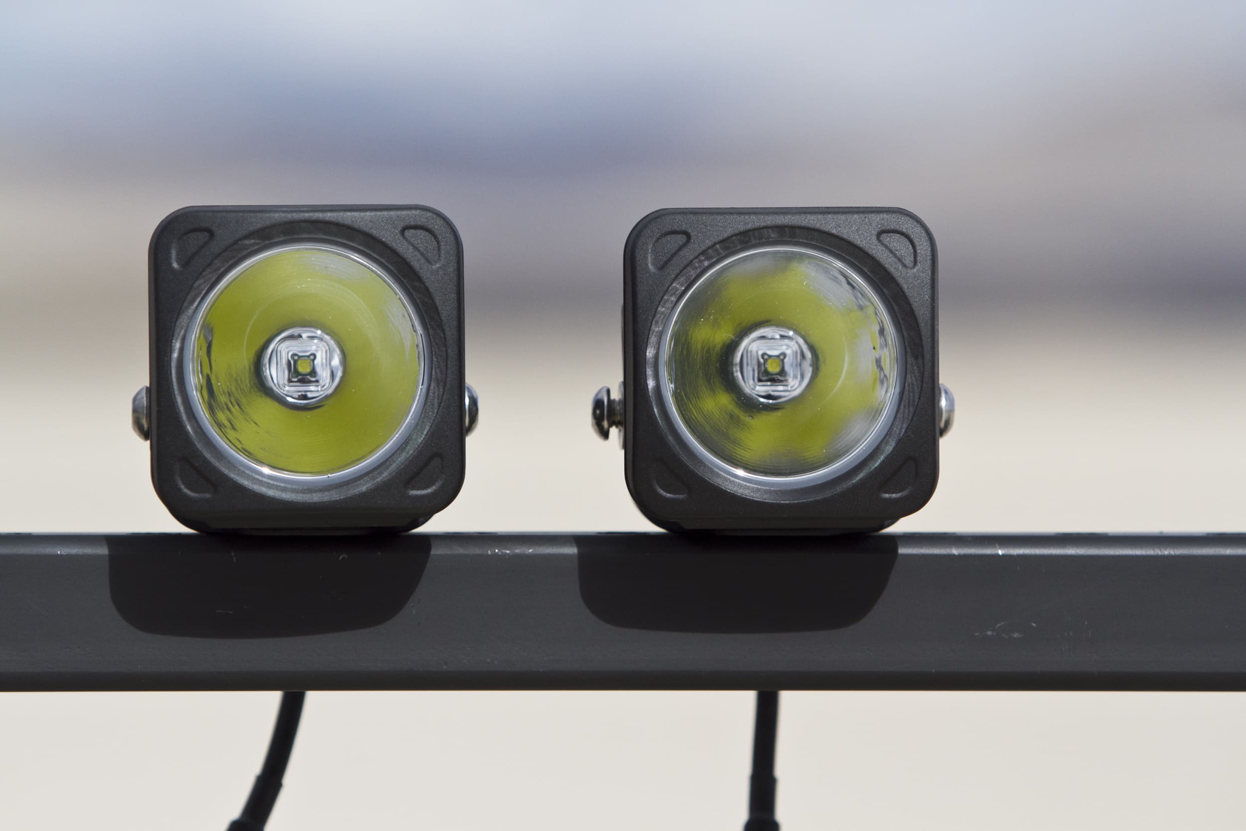 Two square LED driving lights mounted side by side on a horizontal metal bar, with a blurred outdoor background.