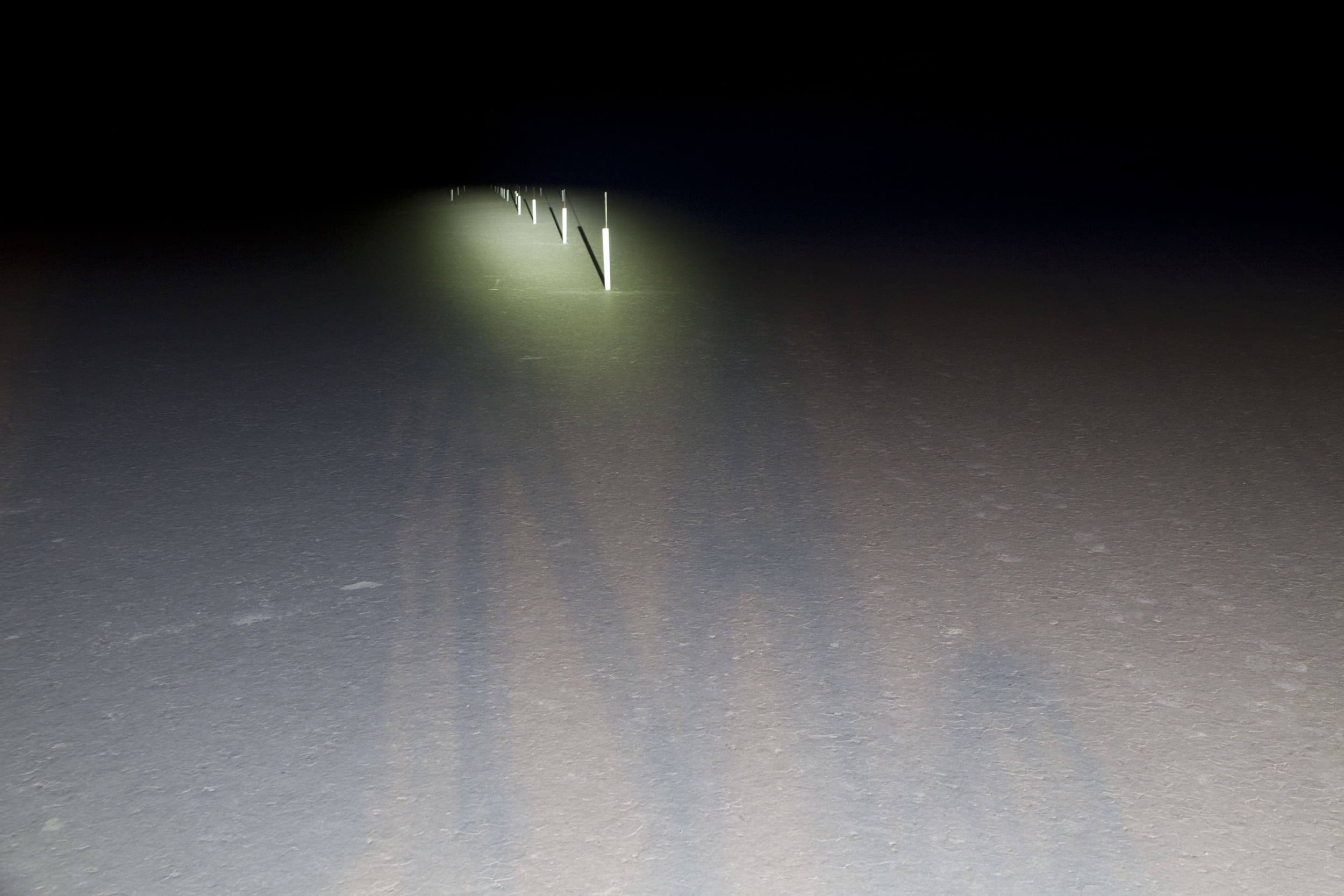 A dark road at night is illuminated by driving lights, casting long shadows on the pavement. Several road markers or posts are faintly visible in the distance within the beam of light.
