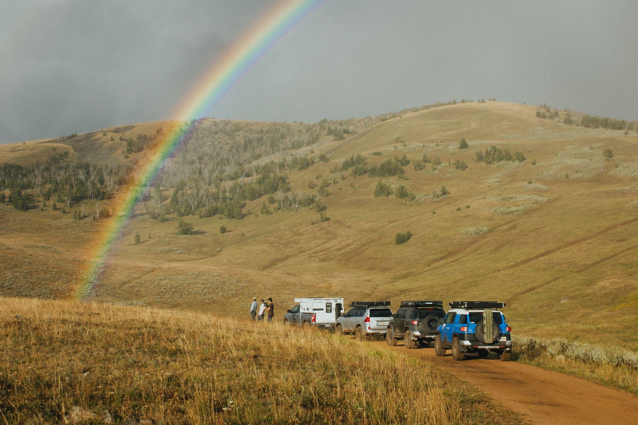 Several vehicles are parked on a dirt road in the grassy, hilly landscape of the Gravelly Range, Montana, with a rainbow arcing across a cloudy sky. People stand near the cars, enjoying the scenic view during their 36 Hours adventure.