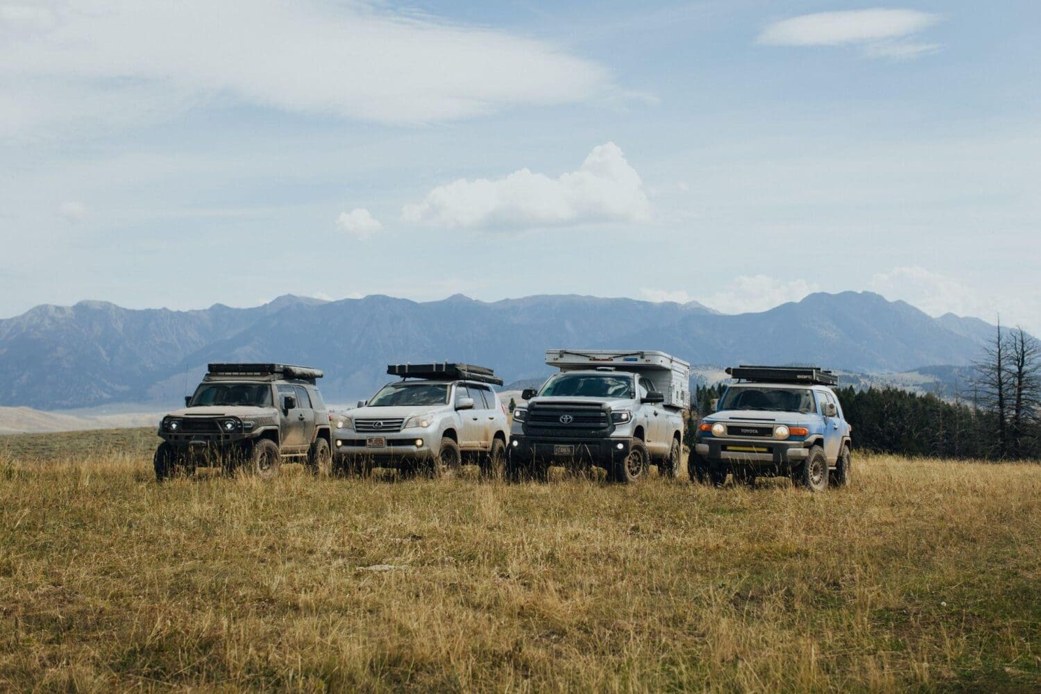 Four off-road vehicles with rooftop tents are parked on grassy terrain in the Gravelly Range, Montana, with mountains and a partly cloudy sky in the background, promising adventure for 36 hours beneath the open sky.