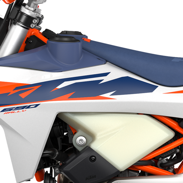Close-up of a 2027 KTM 690 Rally motorcycle showing part of the blue seat, orange and navy graphics, fuel tank, white fairing, and exposed engine components.