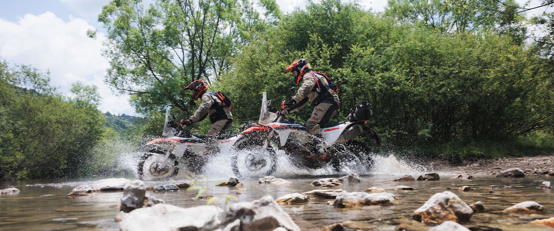 Two people in protective gear ride 2027 KTM 690 Rally dirt bikes through a shallow, rocky stream, splashing water. Trees and greenery surround them under a partly cloudy sky.