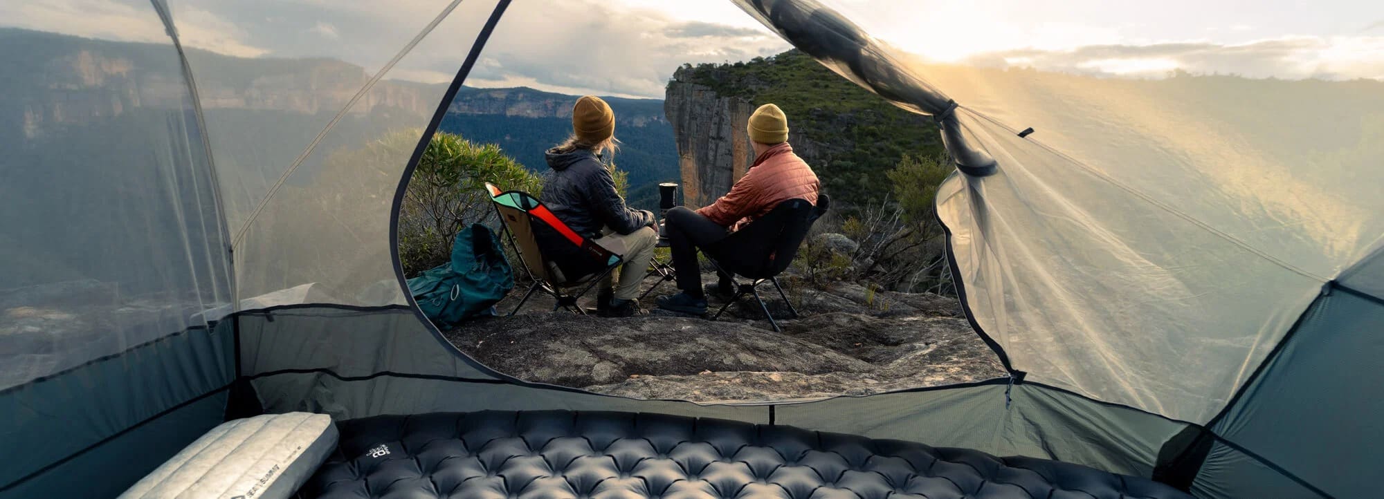 View from inside a tent showing two people in chairs, wearing jackets and hats, sitting near a cliff’s edge and looking at a scenic mountain landscape at sunset. An air mattress is visible in the tent foreground, capturing the quintessential Overland News moment of adventure and outdoor serenity.