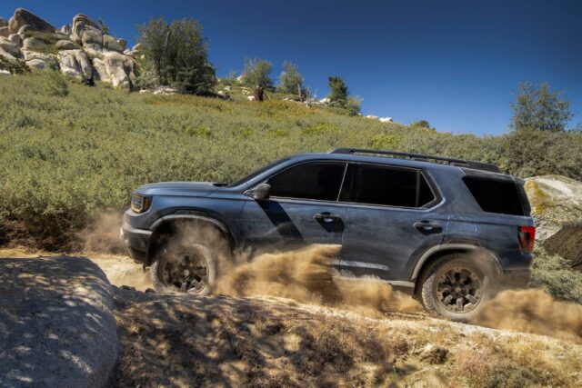A dark blue SUV drives off-road on a dirt trail, kicking up dust and sand, with rocks, bushes, and trees under a clear blue sky in the background—a scene straight out of Overland News.