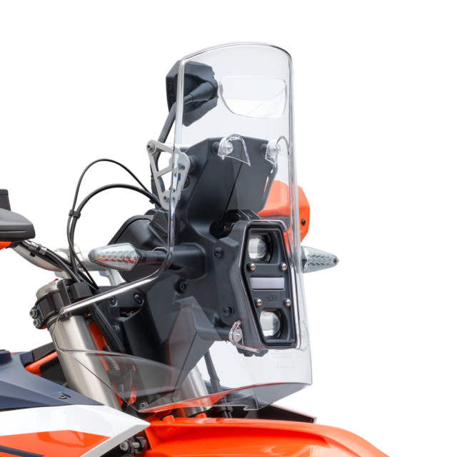 Close-up of a 2027 KTM 690 Rally’s front, showing a clear windscreen, headlight, handlebars, and turn signals, with orange and black bodywork visible against a plain white background.