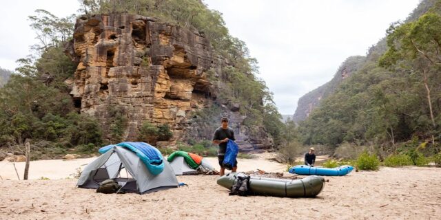 Two people set up tents and packrafts on a sandy riverbank surrounded by trees and tall rocky cliffs under an overcast sky, capturing the spirit of adventure highlighted in Overland News.